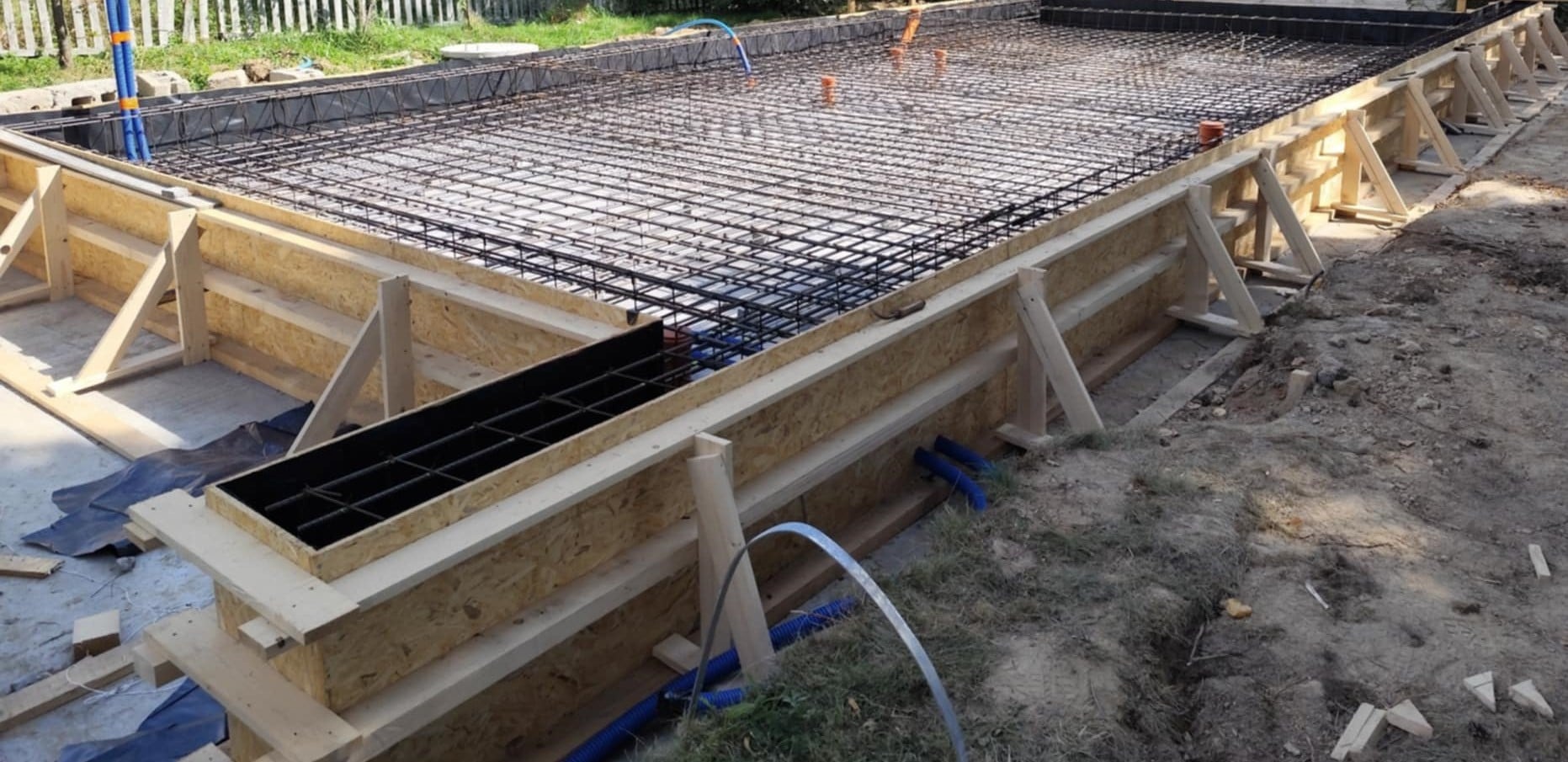 Zbrojenie fundamentów pod budynek z szalunkiem drewnianym, widoczne wystające rury instalacyjne. Siatka zbrojeniowa ułożona, gotowa do zalania betonem.