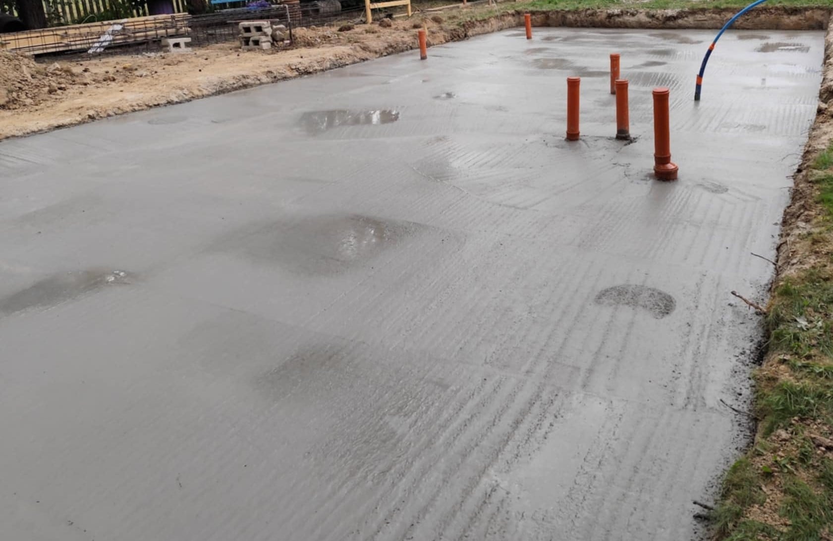 Zalany fundament z wystającymi rurami kanalizacyjnymi. Świeży beton z drobnymi smugami i plamami wody. Widoczne przygotowania do dalszych prac budowlanych.
