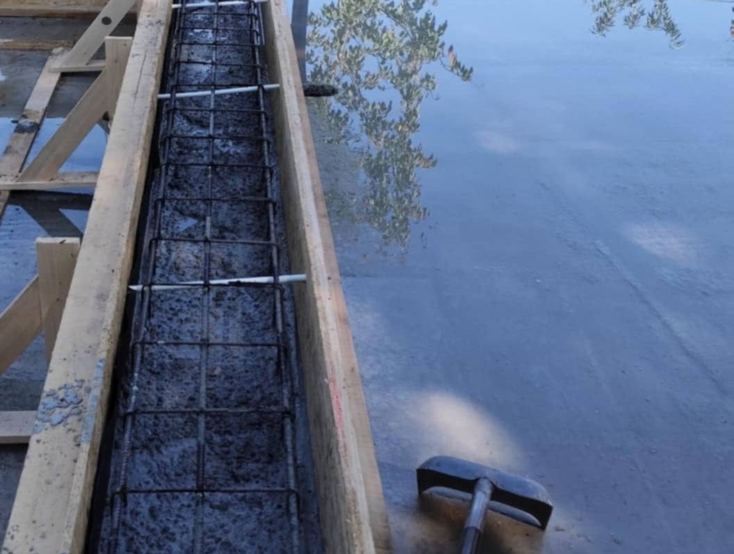 Zbrojenie w szalunku drewnianym, przygotowane do zalania betonem. Widoczne odbicie drzewa w mokrej powierzchni obok i narzędzie budowlane.