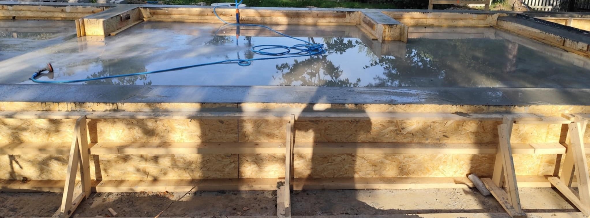 Zalany betonem fundament z drewnianym szalunkiem, niebieski wąż leży na betonie. Widoczne odbicie nieba w mokrej powierzchni betonu.