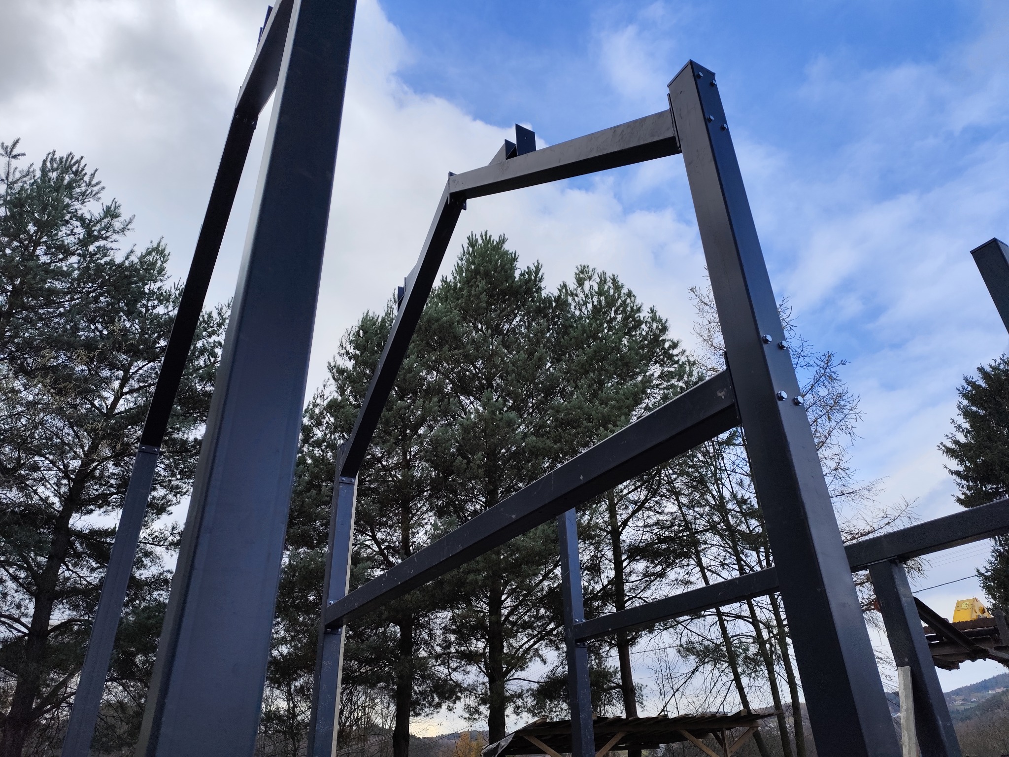 Metalowa konstrukcja szkieletowa w trakcie montażu, widok z dołu na tle drzew i błękitnego nieba z chmurami. Elementy skręcane śrubami.