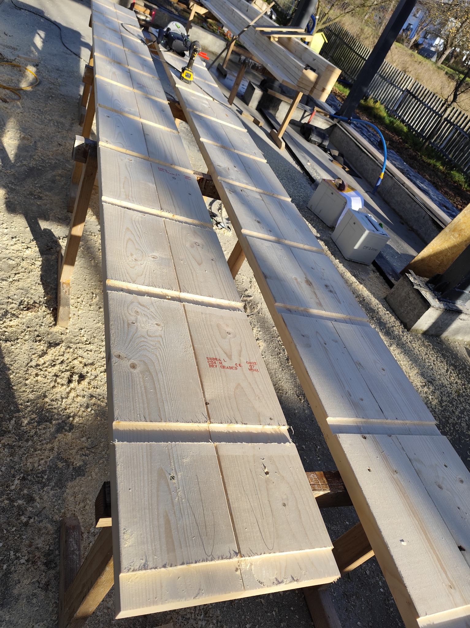 Długie, jasne deski na drewnianych kozłach, przygotowywane do montażu. W tle narzędzia i elementy konstrukcyjne. Widoczne ślady obróbki drewna.