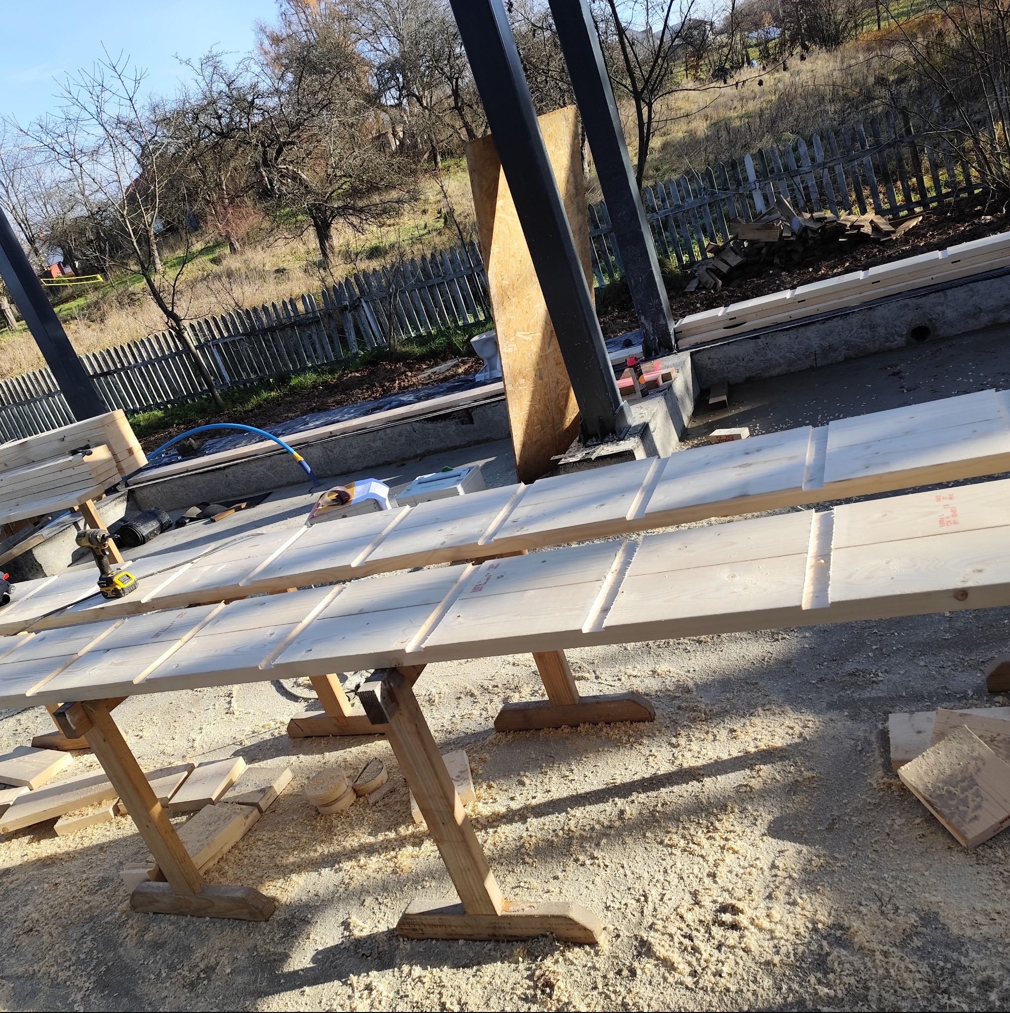 Montaż drewnianych elementów konstrukcyjnych na placu budowy. Widoczne narzędzia i drewniane podpory. W tle ogrodzenie i zieleń.