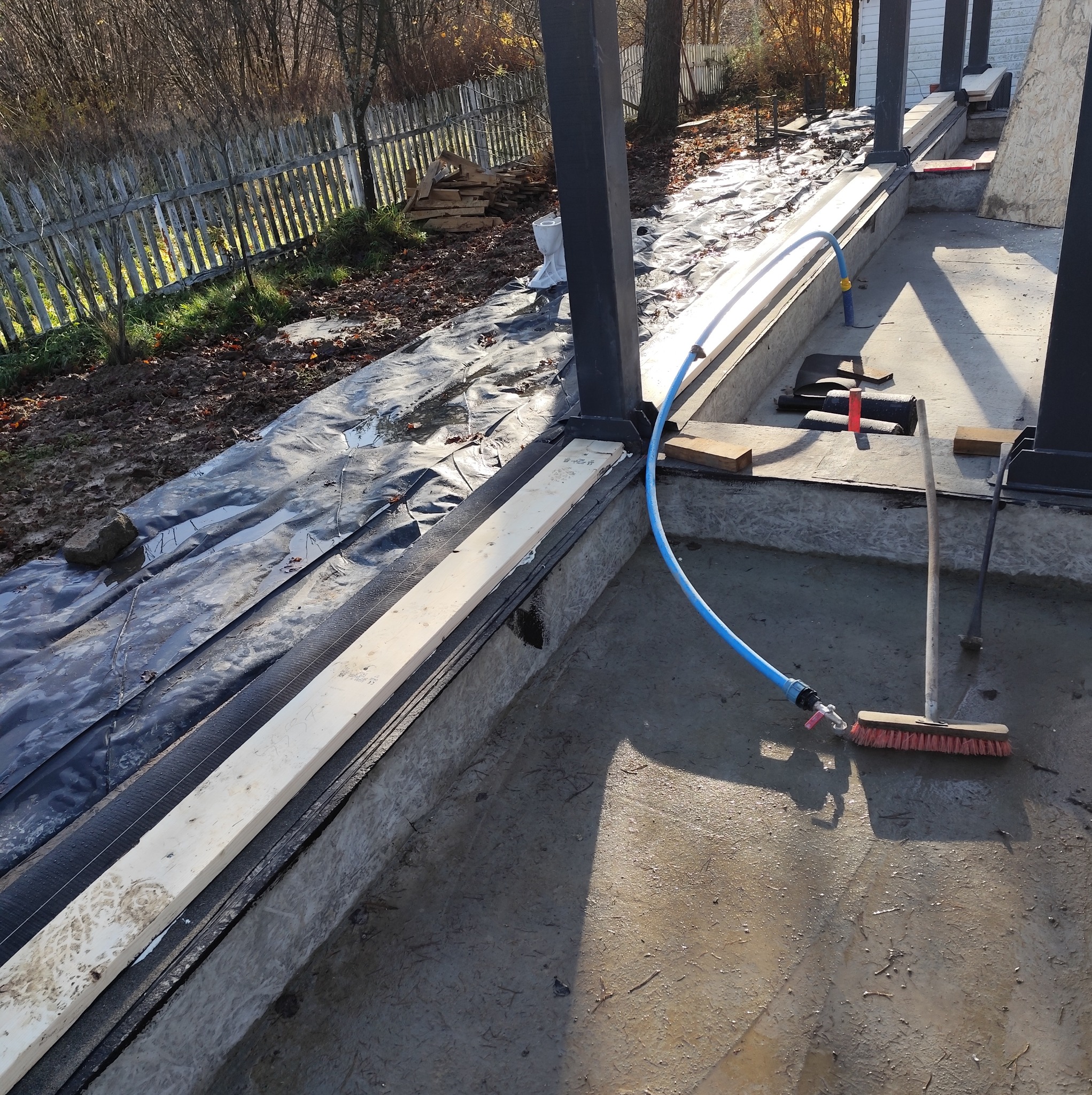 Wylewka fundamentowa z hydroizolacją i deskowaniem, przygotowana pod budowę. Widoczny wąż z wodą i narzędzia budowlane. W tle ogrodzenie i drzewa.
