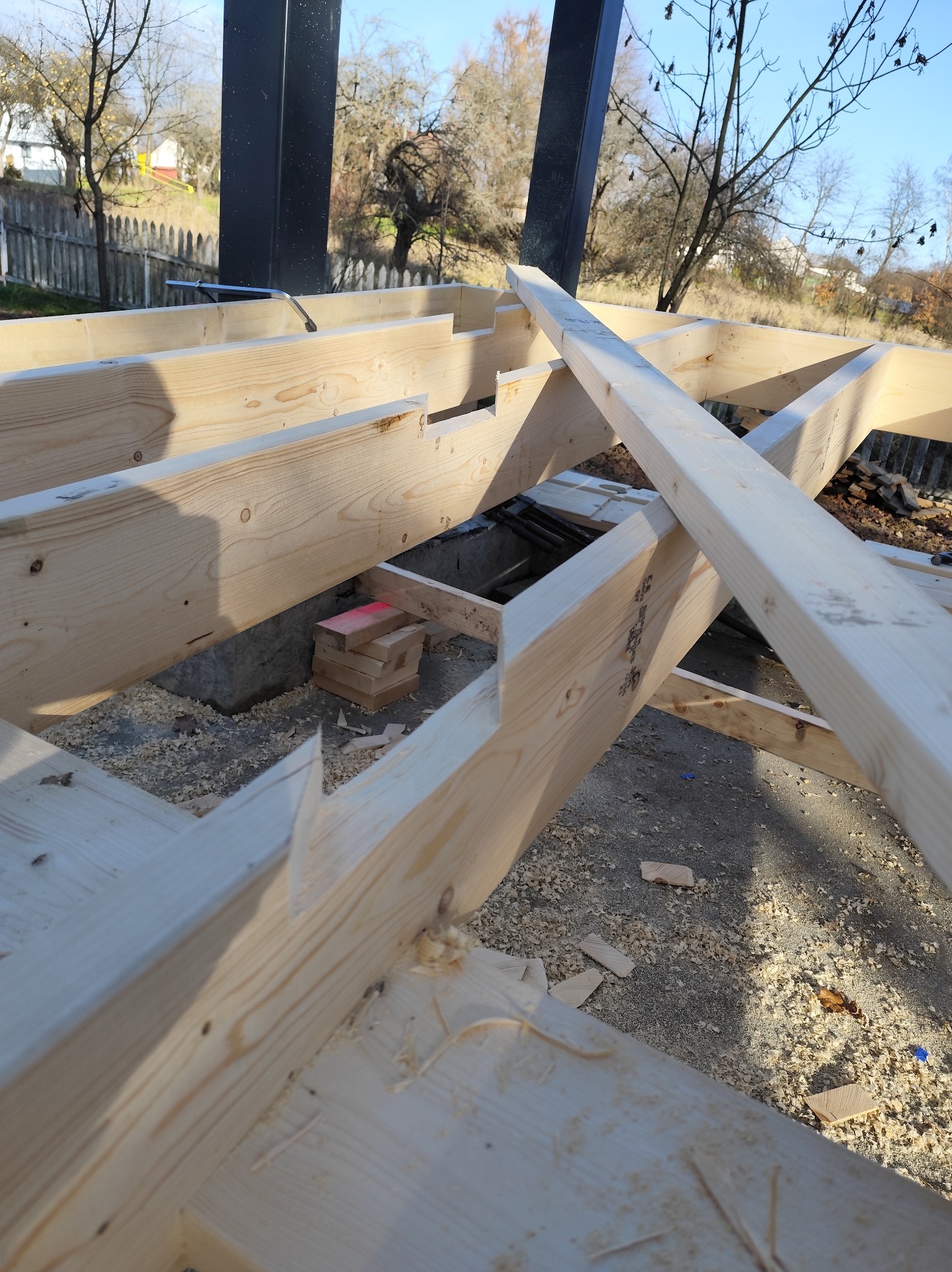 Konstrukcja szkieletowa z jasnego drewna w trakcie montażu, widoczne elementy fundamentu i słupy nośne. W tle drzewa i ogrodzenie.