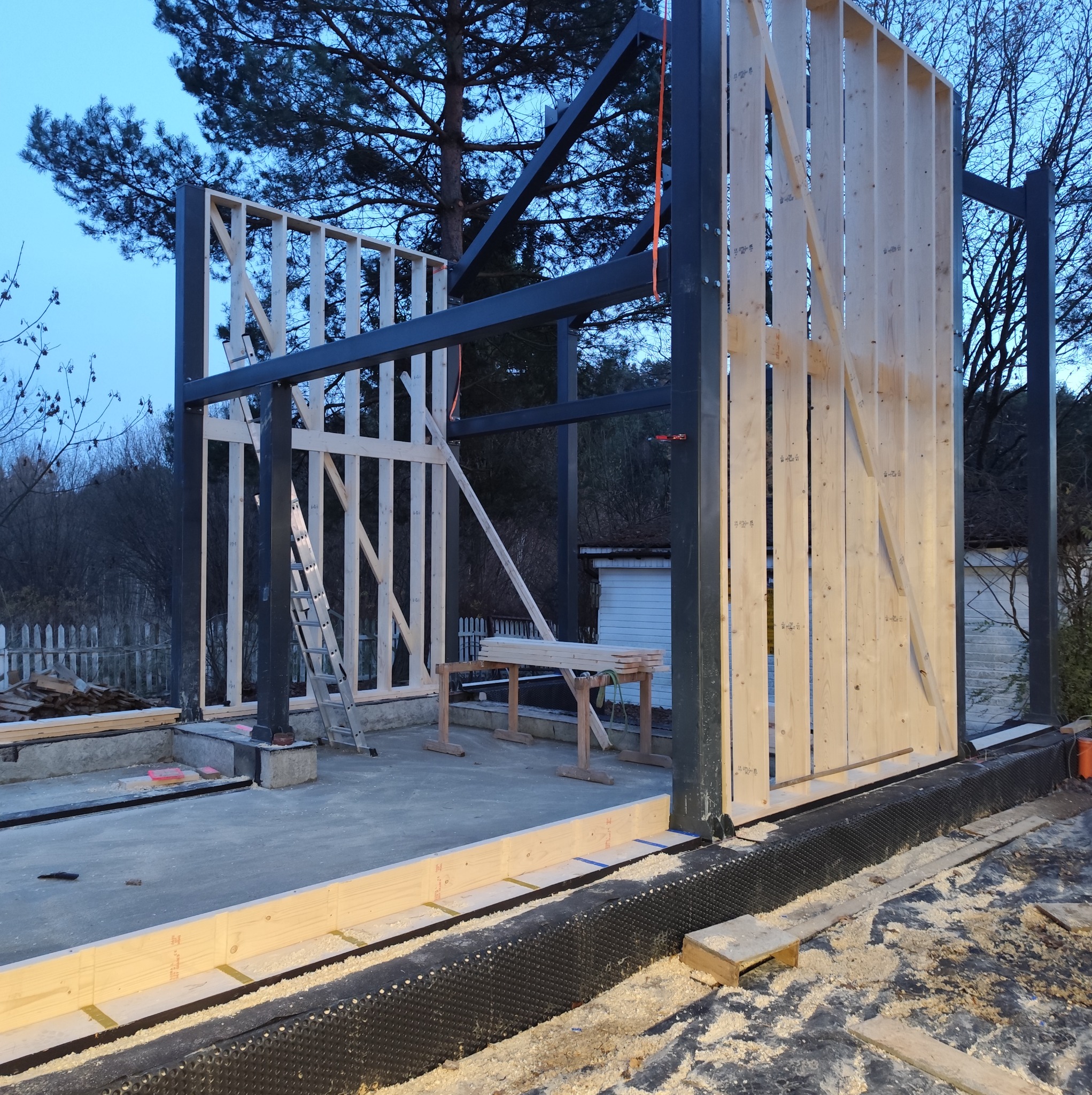 Wznoszenie konstrukcji domu szkieletowego z elementami stalowymi. Widoczny fundament, ściany w trakcie budowy, drewniane elementy i stalowe słupy. W tle drzewa i budynek.