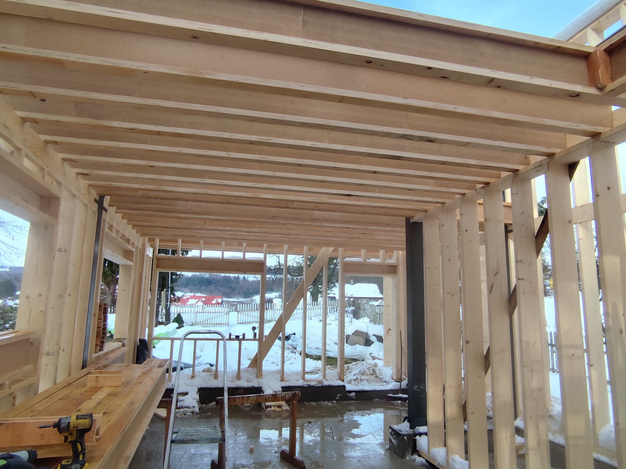 Wnętrze domu szkieletowego w trakcie budowy. Widoczne drewniane belki stropowe i ściany. Na zewnątrz zimowy krajobraz z ośnieżonymi domami. Wnętrze w stanie surowym.