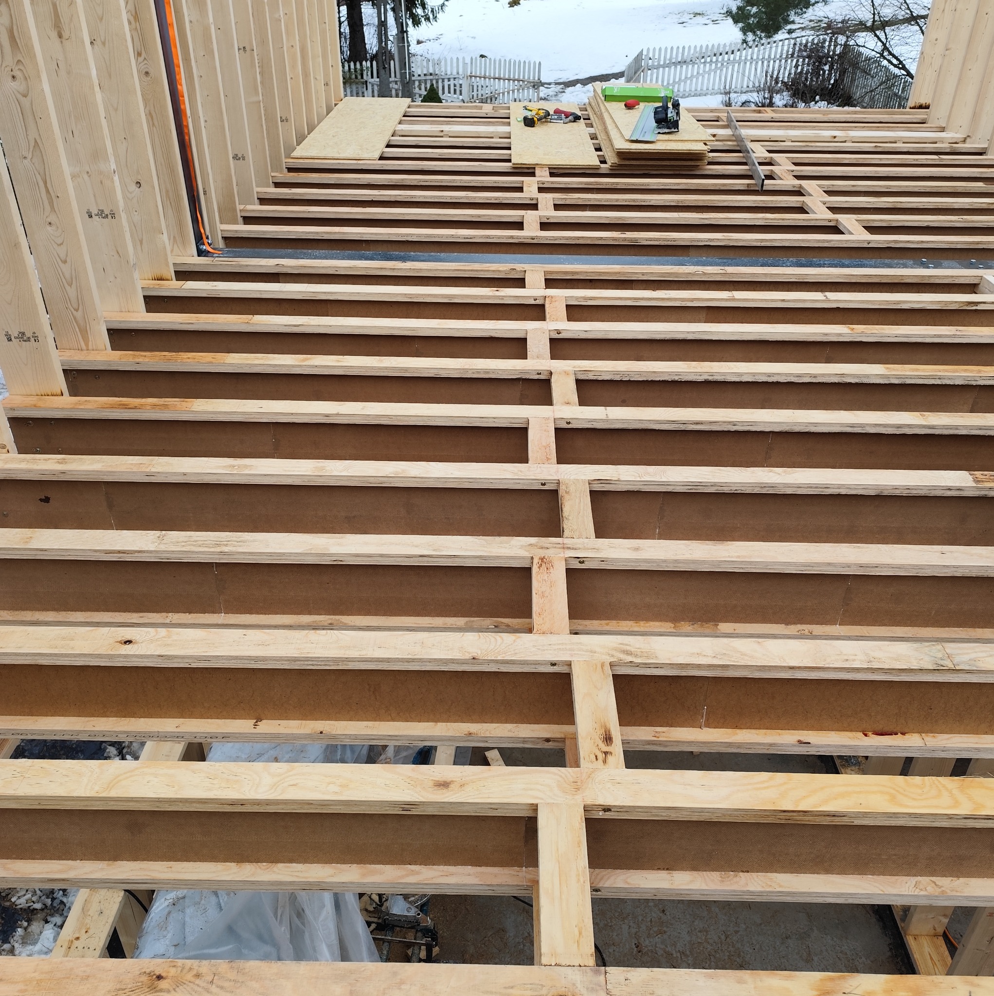 Konstrukcja szkieletowa domu w trakcie budowy. Widoczne drewniane belki stropowe i ściany, narzędzia na górze. W tle ośnieżony teren.