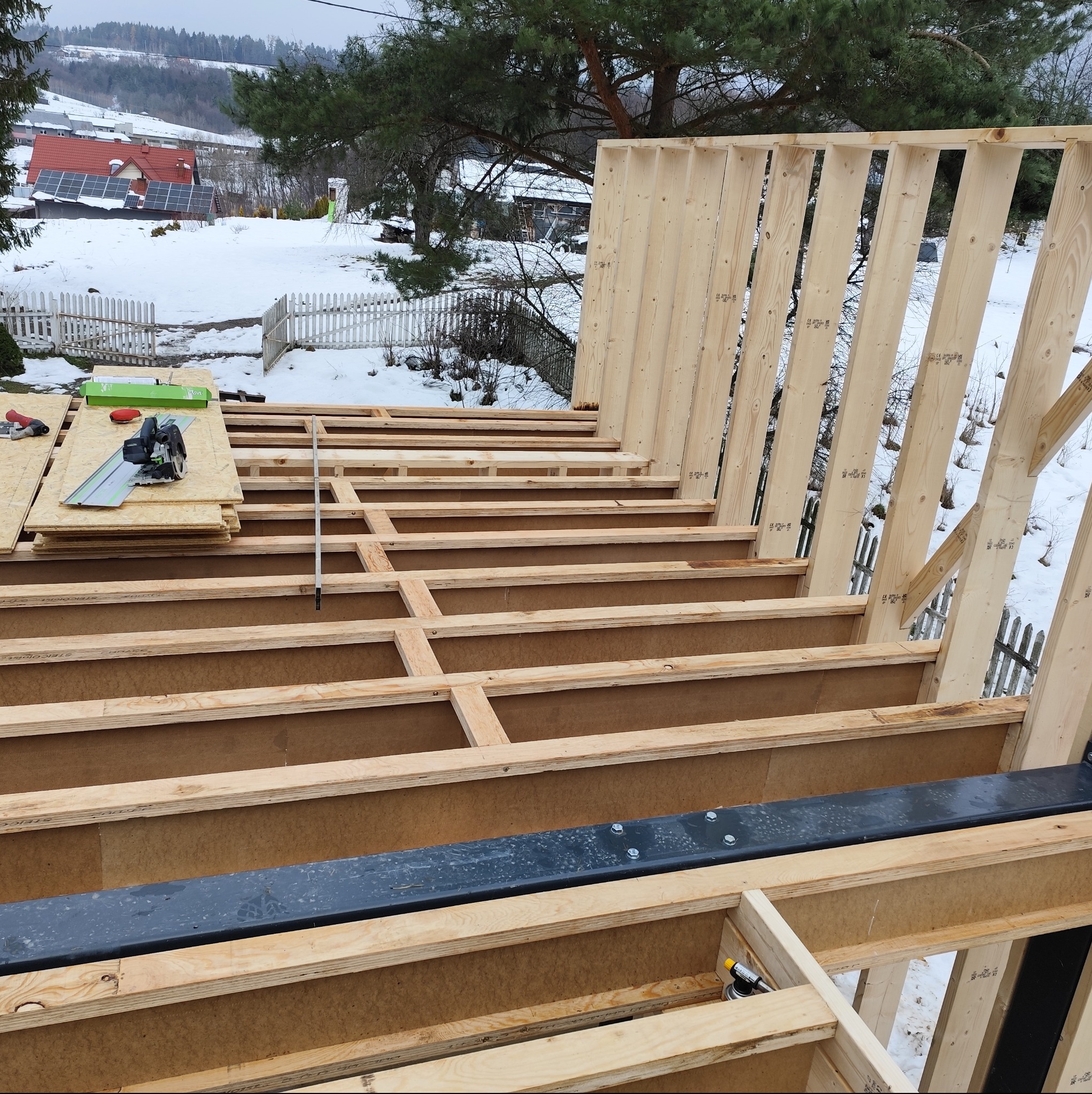 Konstrukcja szkieletowa domu w trakcie budowy, widoczne drewniane elementy nośne i narzędzia na tle zimowego krajobrazu. Ujęcie z góry, detale łączeń.