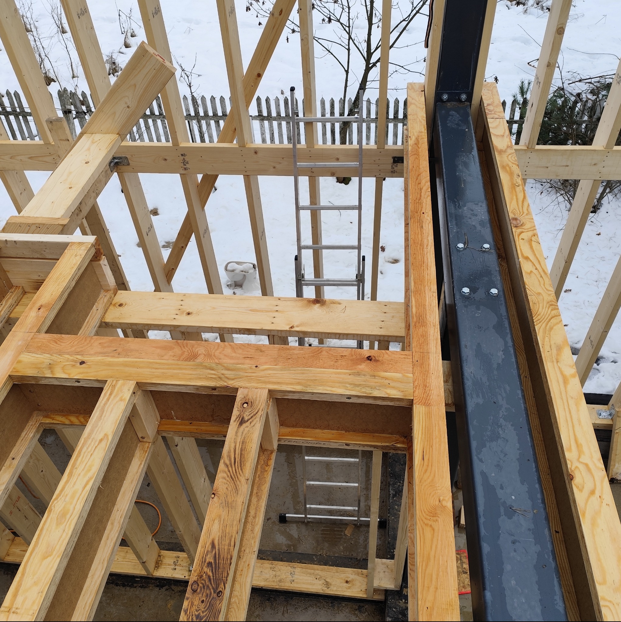 Konstrukcja drewniana dachu w trakcie budowy, widok z góry. Elementy złączone, widoczna drabina i zimowy krajobraz w tle. Stalowy słup wzmacniający konstrukcję.