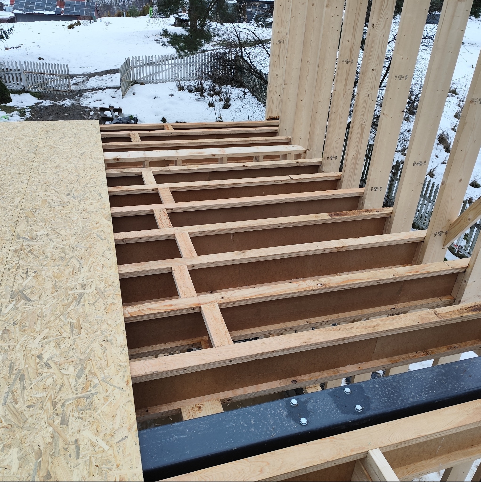 Konstrukcja szkieletowa w trakcie budowy: drewniane belki stropowe i pionowe słupy, częściowo pokryte płytą OSB. W tle zimowy krajobraz.