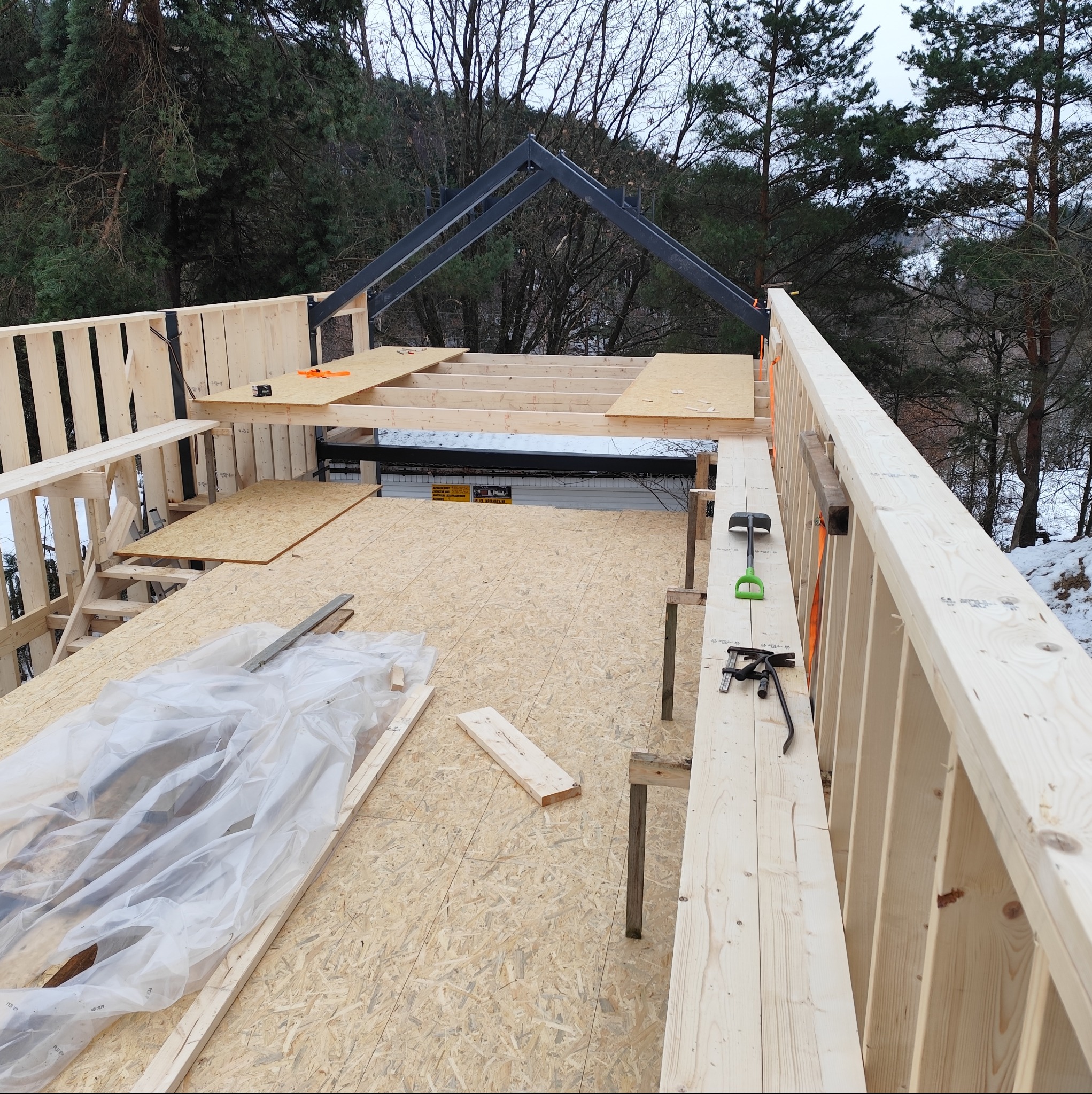Konstrukcja domu w trakcie budowy: drewniane ściany, płyty OSB na podłodze, stalowa konstrukcja dachu. Widoczne narzędzia i elementy montażowe. Otoczenie leśne.