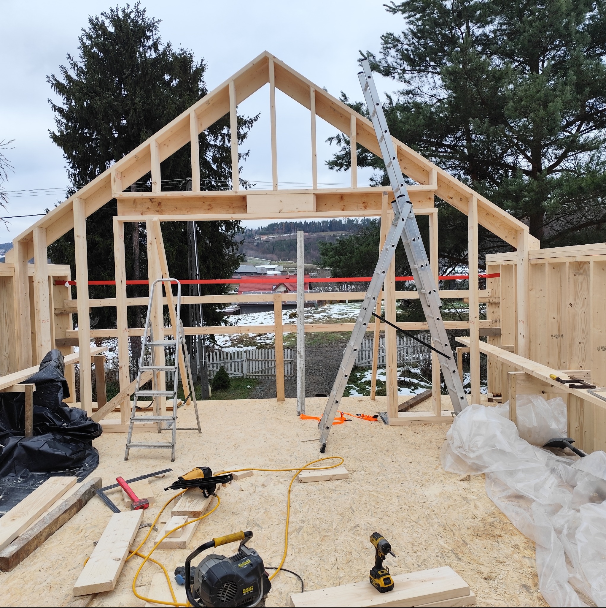 Wnętrze domu szkieletowego w trakcie budowy. Widoczne drewniane ramy konstrukcyjne, narzędzia i drabiny. W tle krajobraz z elementami zimowego pejzażu.