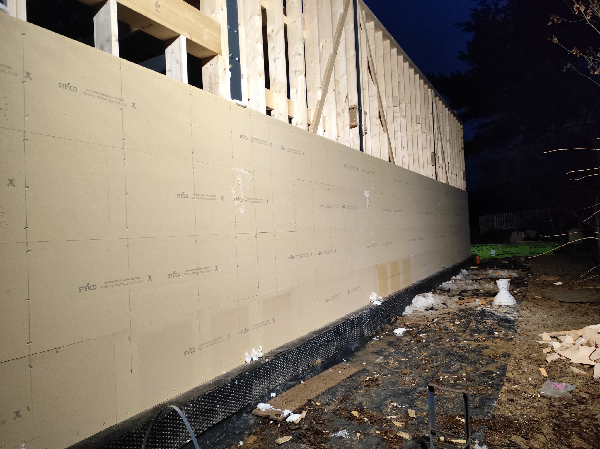 Budowa domu szkieletowego: widok na ścianę z płyt Steico, z widoczną konstrukcją drewnianą. Ujęcie z zewnątrz, w tle ciemne niebo i drzewa. Widoczne detale montażu.