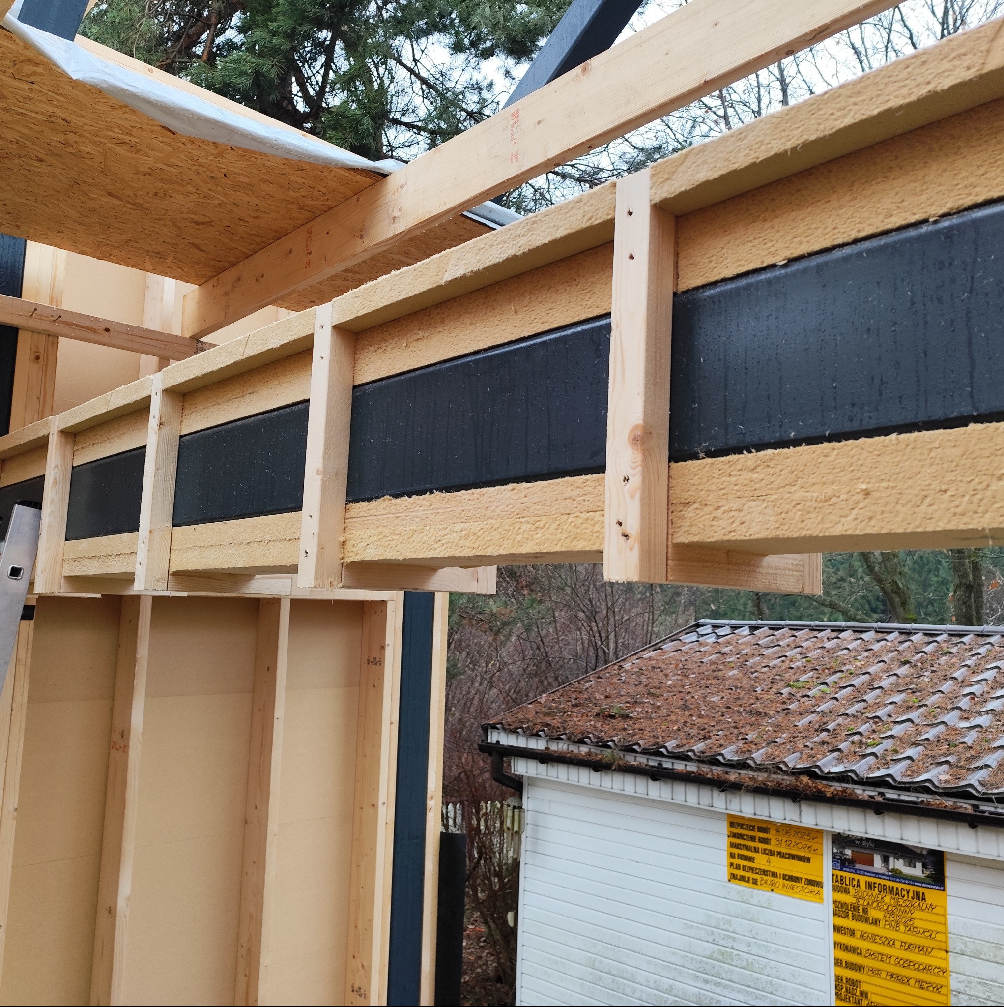 Wnętrze w trakcie budowy domu szkieletowego: drewniana konstrukcja z izolacją, widoczne elementy dachu i ściany. W tle budynek z tablicą informacyjną budowy.