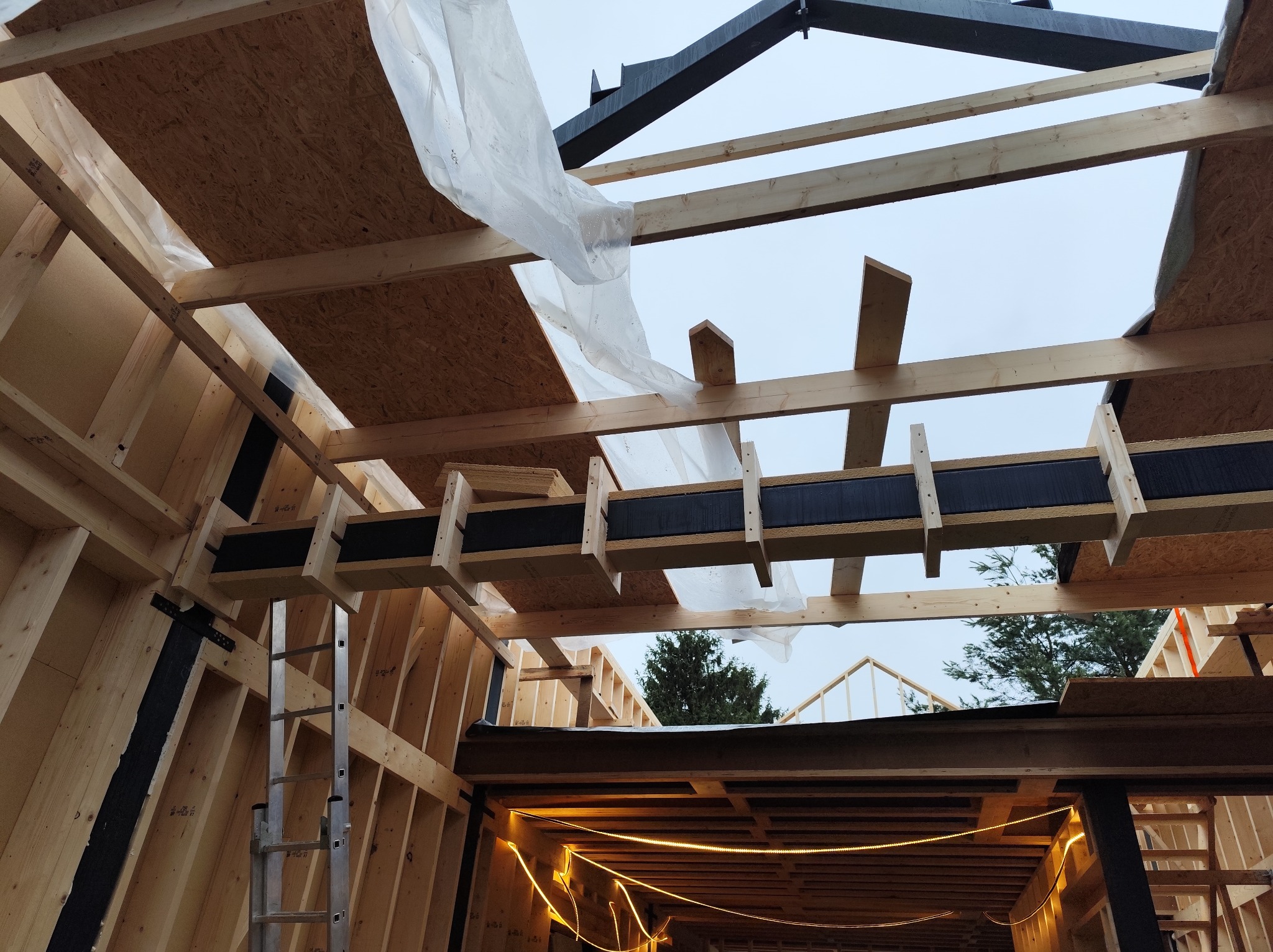 Wnętrze w trakcie budowy domu szkieletowego. Widoczne drewniane elementy konstrukcyjne, płyty OSB, folia ochronna i drabina. Ujęcie z dołu, w tle niebo.
