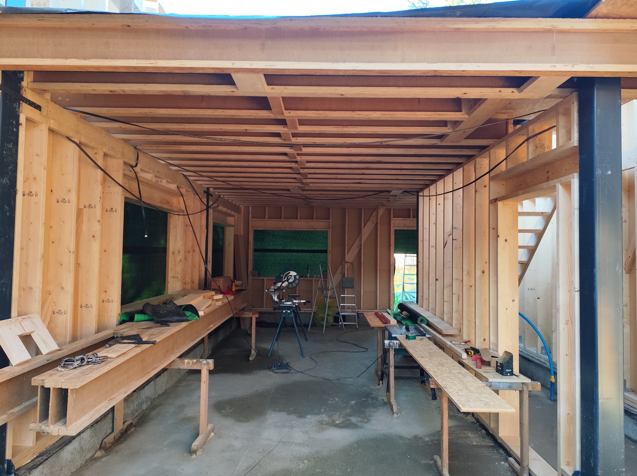 Wnętrze surowego domu szkieletowego w trakcie budowy. Widoczne drewniane elementy konstrukcyjne, narzędzia i stanowiska pracy. Surowy beton na podłodze.