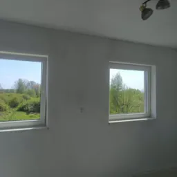 Pok&oacute;j po gładziowaniu ścian, widoczne dwa okna z widokiem na zieleń, surowe wykończenie podłogi, gotowe do malowania.
