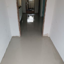 DAW-TYNK - Świeżo wylana, mokra, jasnoszara posadzka w trakcie schnięcia, widok z góry na korytarz z otwartymi drzwiami do kolejnych pomieszczeń.