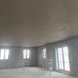 DAW-TYNK - Wnętrze w trakcie prac wykończeniowych: ściany pokryte surowym tynkiem, metalowe rusztowania przy oknach. Widoczne przewody elektryczne na suficie i niebieska rurka przy podłodze.