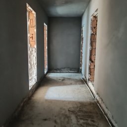 DAW-TYNK - Wnętrze korytarza w trakcie prac wykończeniowych: ściany po tynkowaniu, widoczne fragmenty cegieł w otworach drzwiowych, surowa posadzka.