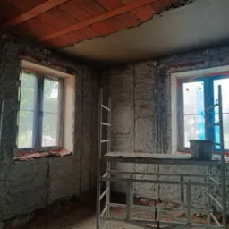Wnętrze w trakcie prac tynkarskich: ściany pokryte surowym tynkiem, widoczne rusztowania i okna zabezpieczone folią. Fragment sufitu z ceglanym wykończeniem.