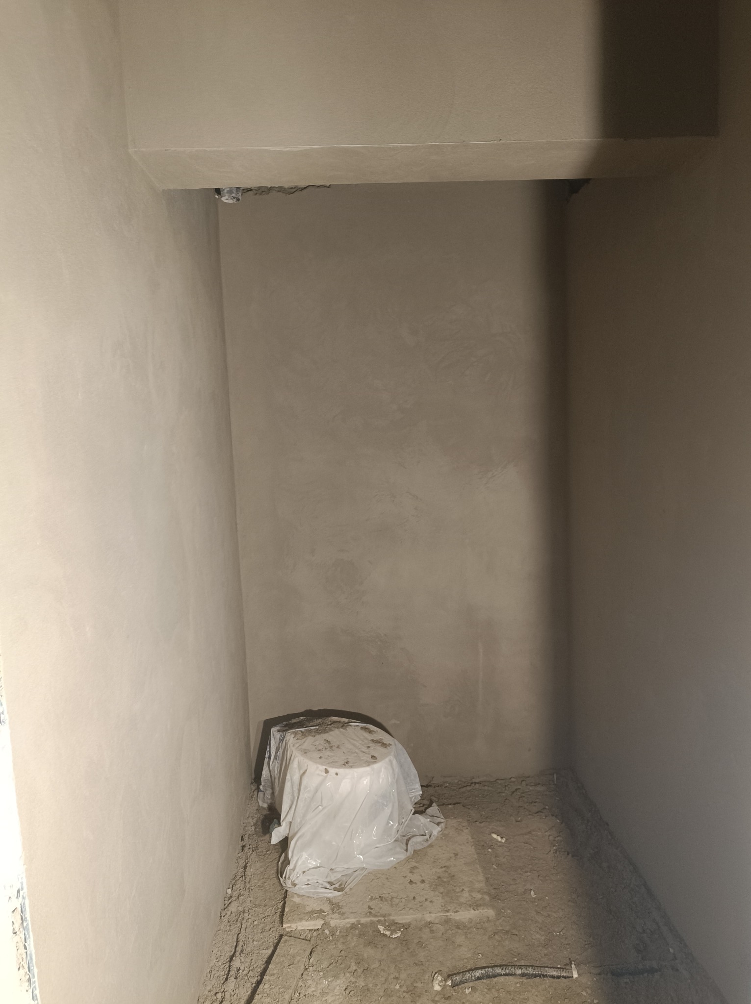 Wnętrze pomieszczenia w trakcie prac wykończeniowych, ściany pokryte świeżo nałożoną, jasną gładzią, podłoga częściowo zakryta gruzem i fragmentami materiałów budowlanych, element instalacji...