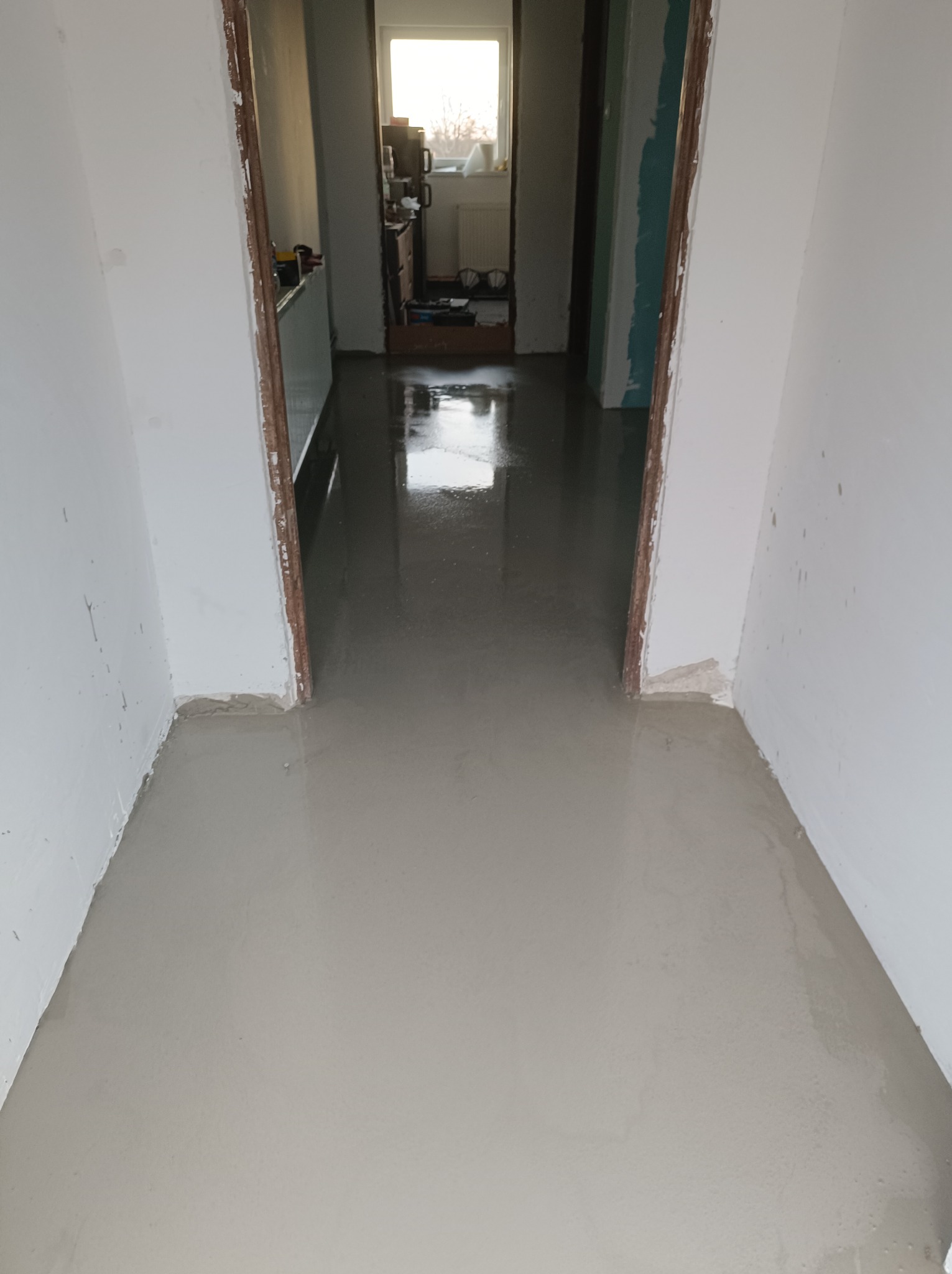 Świeżo wylana, mokra, jasnoszara posadzka w trakcie schnięcia, widok z góry na korytarz z otwartymi drzwiami do kolejnych pomieszczeń.