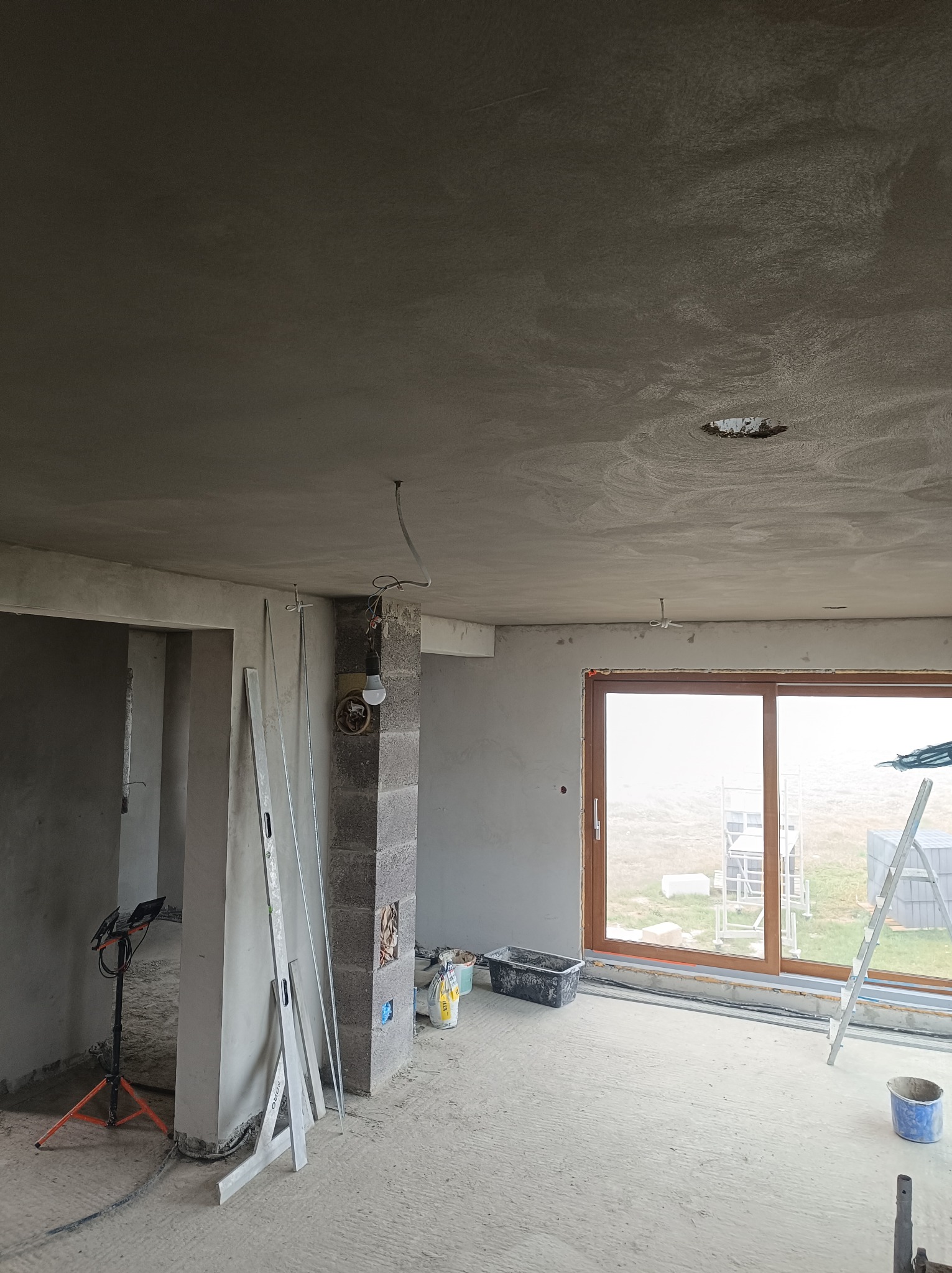 Surowe wnętrze w trakcie remontu: świeżo położona warstwa szarej zaprawy na suficie i ścianach, widoczne narzędzia - poziomice, lampa na statywie, drabina oparta o okno, oraz worki z zaprawą...