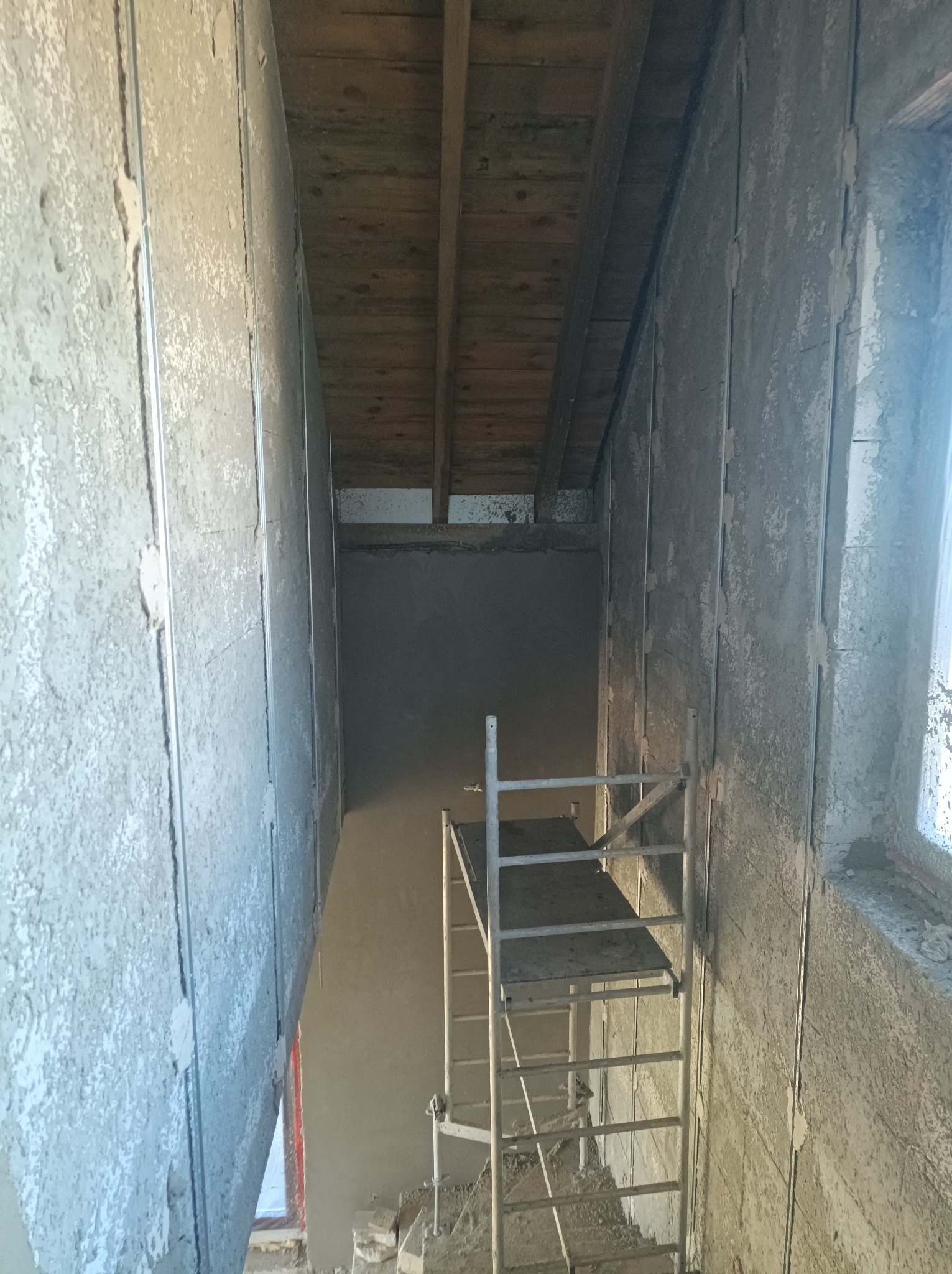 Wnętrze w trakcie remontu: ściany z surowym tynkiem, widoczne metalowe profile, drewniany sufit i drabina aluminiowa w pomieszczeniu z oknem.