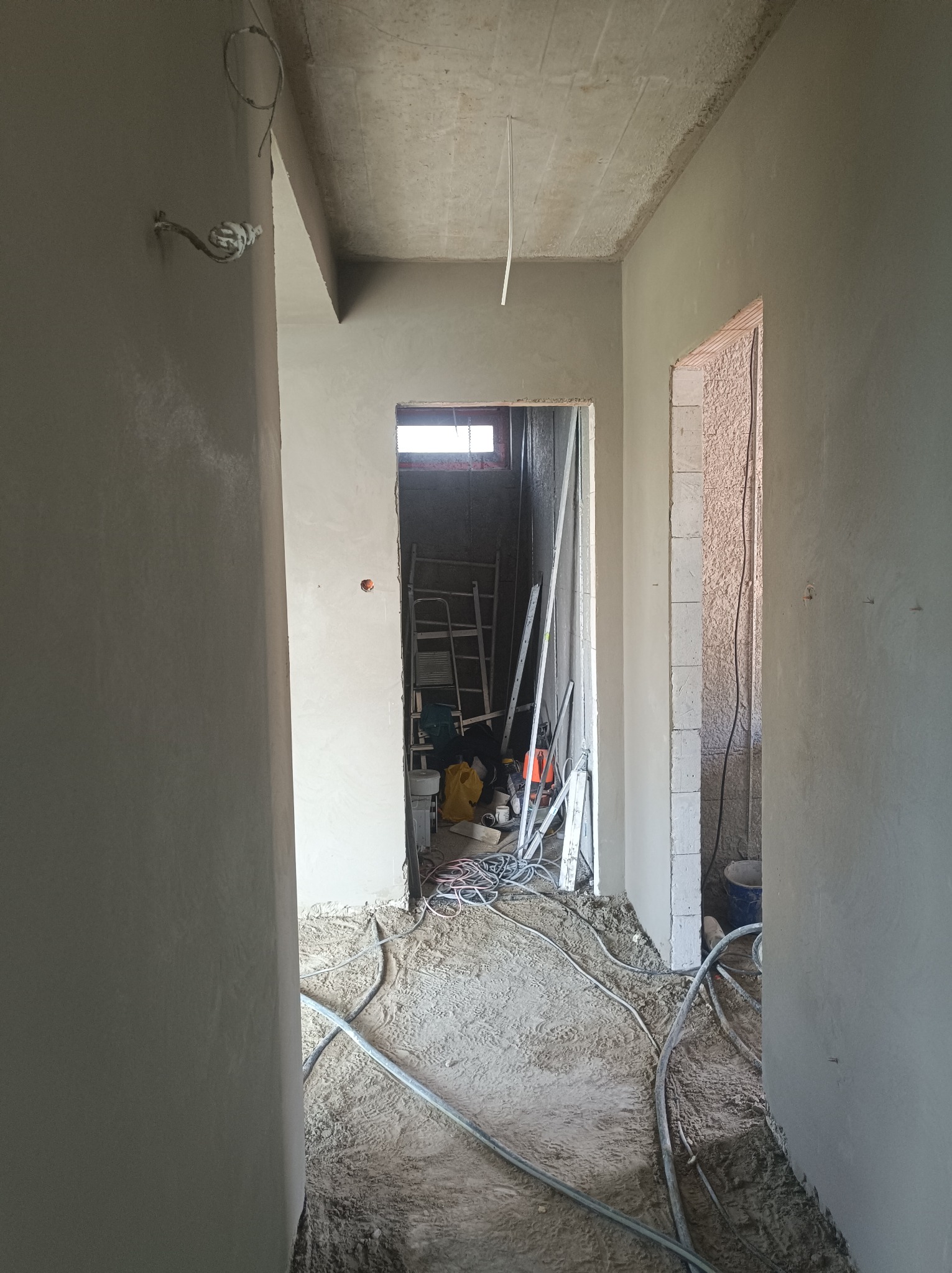Korytarz w trakcie prac wykończeniowych: świeżo nałożone tynki na ścianach, widoczne przewody elektryczne, drabina i narzędzia w tle, podłoga pokryta resztkami materiałów budowlanych.