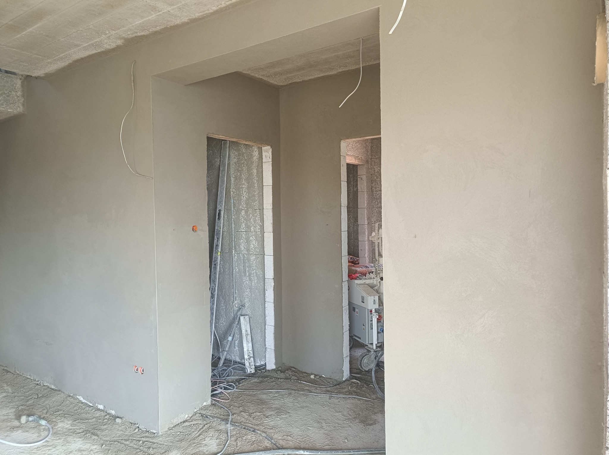 Wnętrze w trakcie prac wykończeniowych; ściany pokryte świeżo nałożonym tynkiem, widoczne przewody elektryczne i maszyna do tynkowania w tle, podłoże zanieczyszczone resztkami materiałów budowlanych.