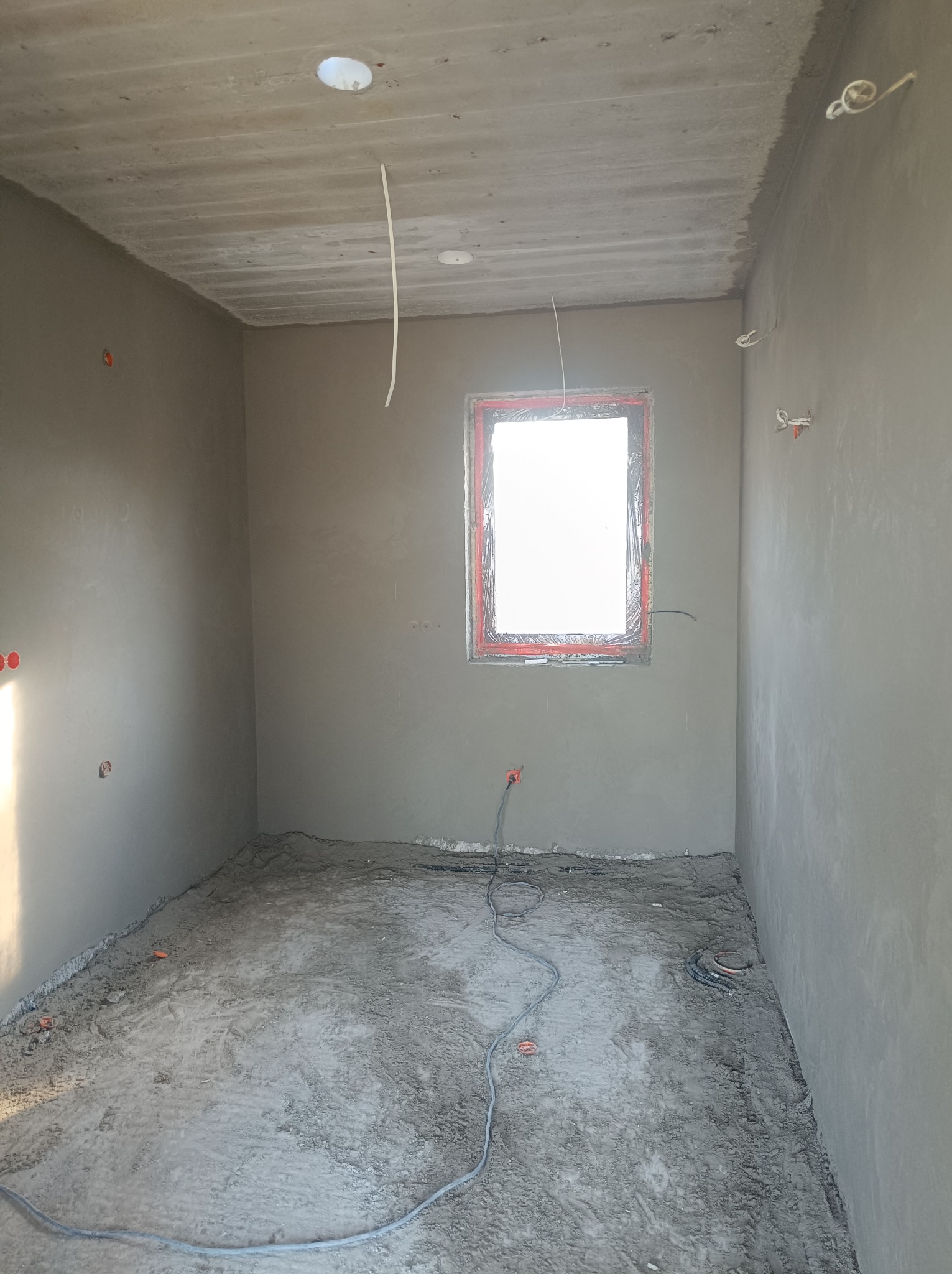 Surowe wnętrze w trakcie prac wykończeniowych: świeżo otynkowane ściany, betonowa wylewka na podłodze, wystające przewody elektryczne i okno zabezpieczone folią.
