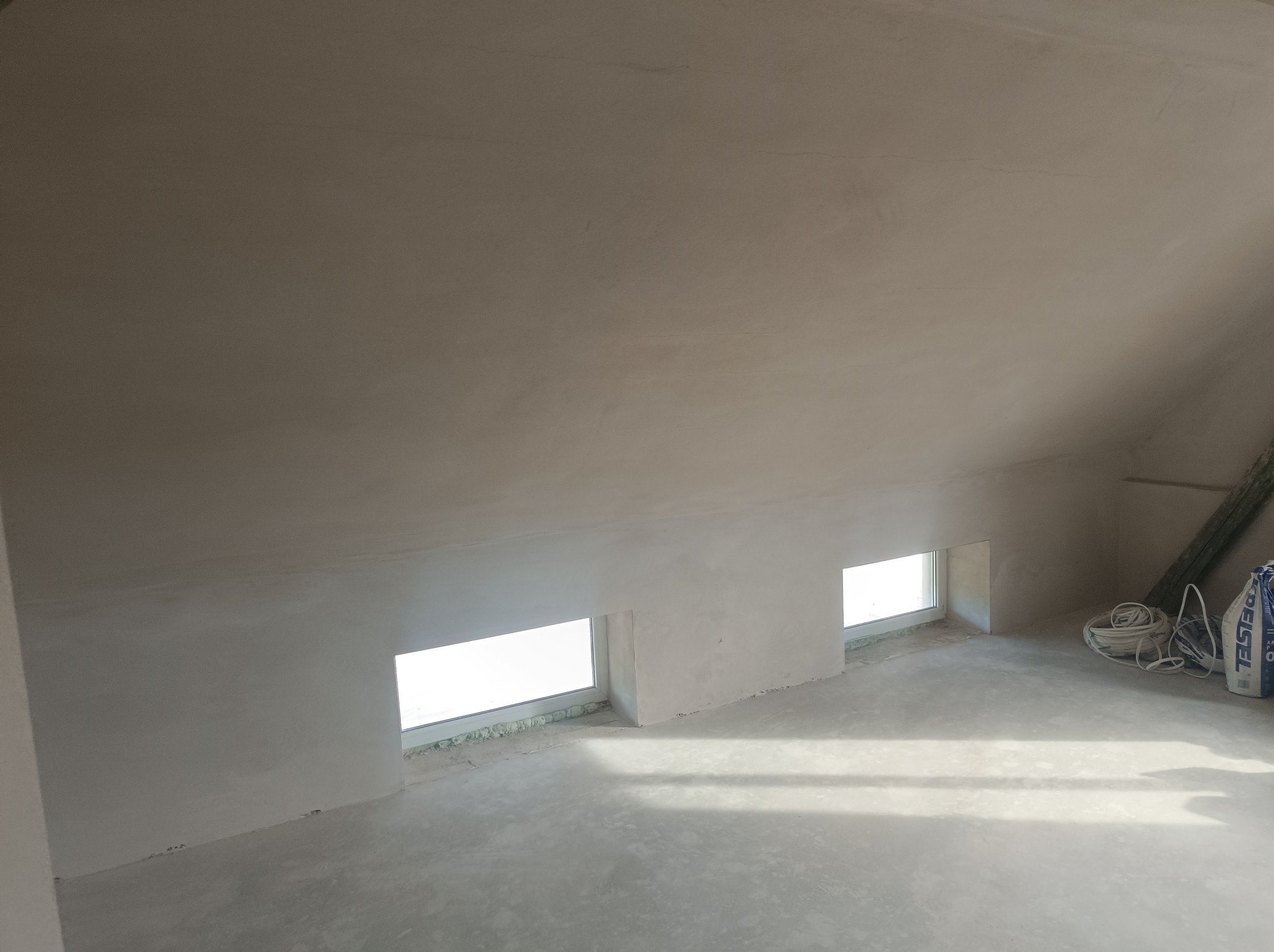 Poddasze po nałożeniu jasnej, gładkiej warstwy wykończeniowej na ścianach i suficie, widoczne dwa małe okna dachowe i wiązka przewodów oraz worek z zaprawą.