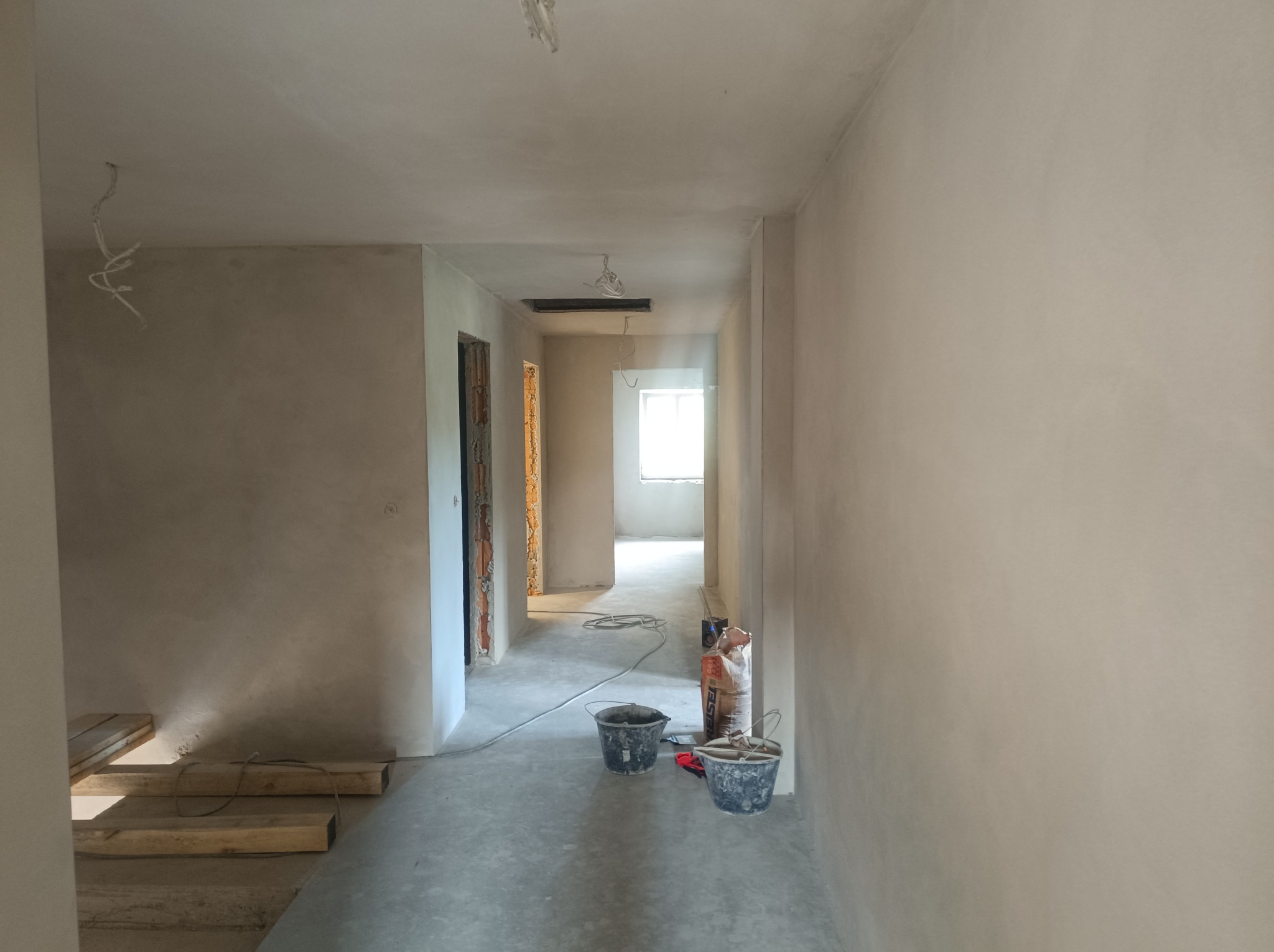 Wnętrze w trakcie prac wykończeniowych: wygładzone ściany w odcieniach beżu, widoczne wiadra, worki z zaprawą i kable wystające z sufitu w długim korytarzu z otwartymi przejściami do kolejnych...