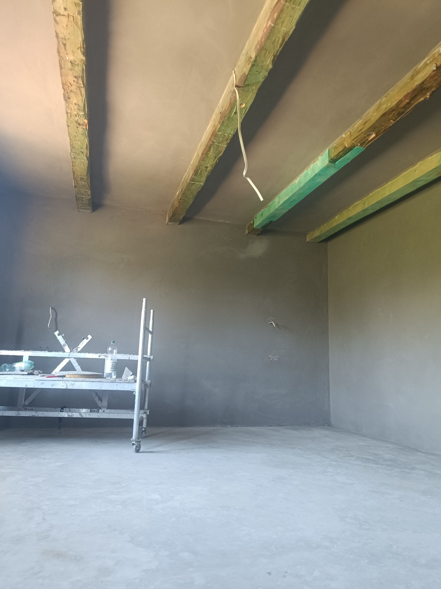 Wnętrze pomieszczenia w trakcie prac wykończeniowych, ściany i sufit pokryte świeżą warstwą szarego tynku, widoczne drewniane belki stropowe, metalowy stół roboczy z narzędziami i butelką wody.
