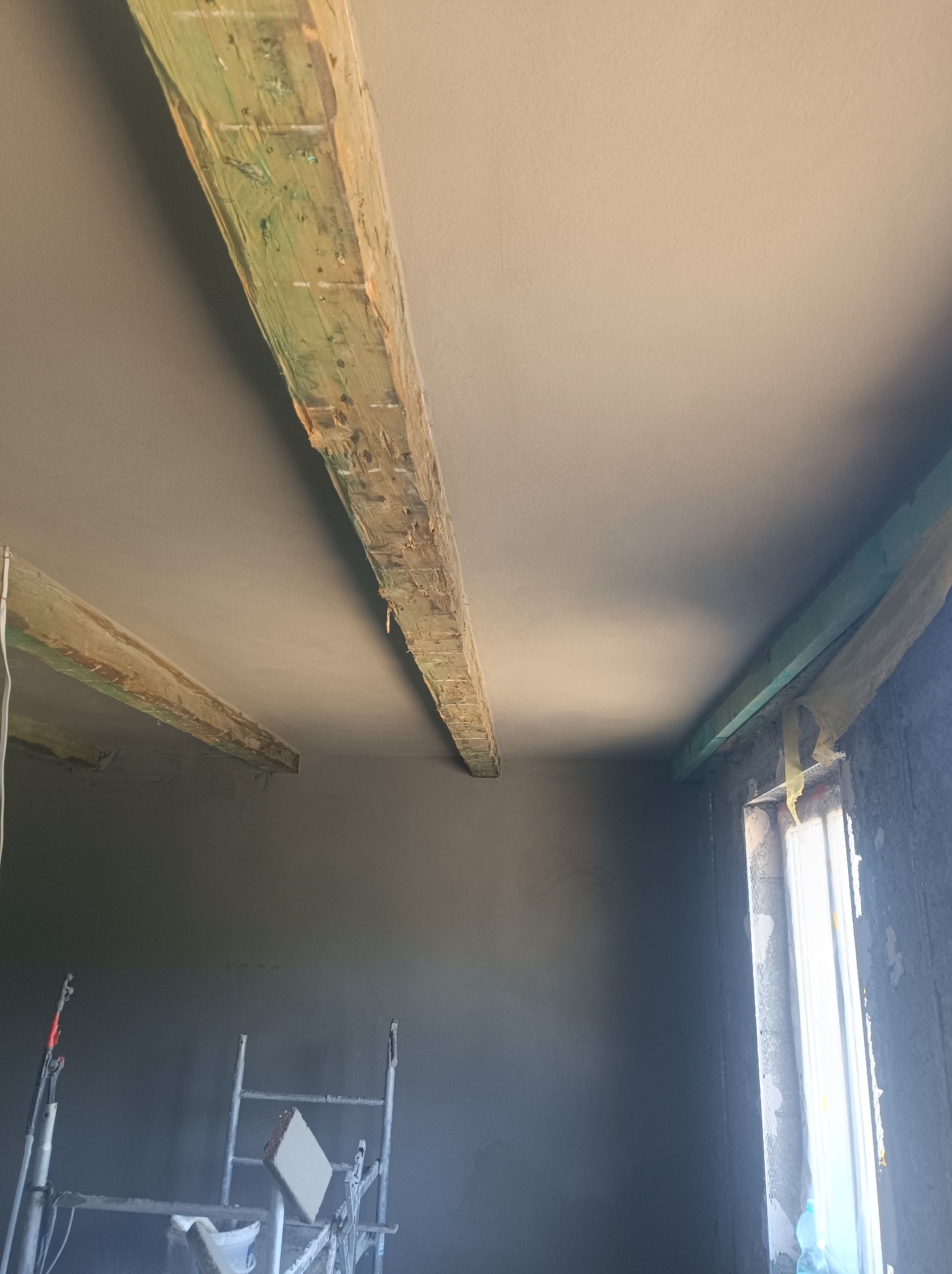 Wnętrze w trakcie prac wykończeniowych: surowa ściana i sufit po nałożeniu warstwy wykończeniowej, widoczne drewniane belki stropowe, rusztowanie malarskie i okno z taśmą ochronną.