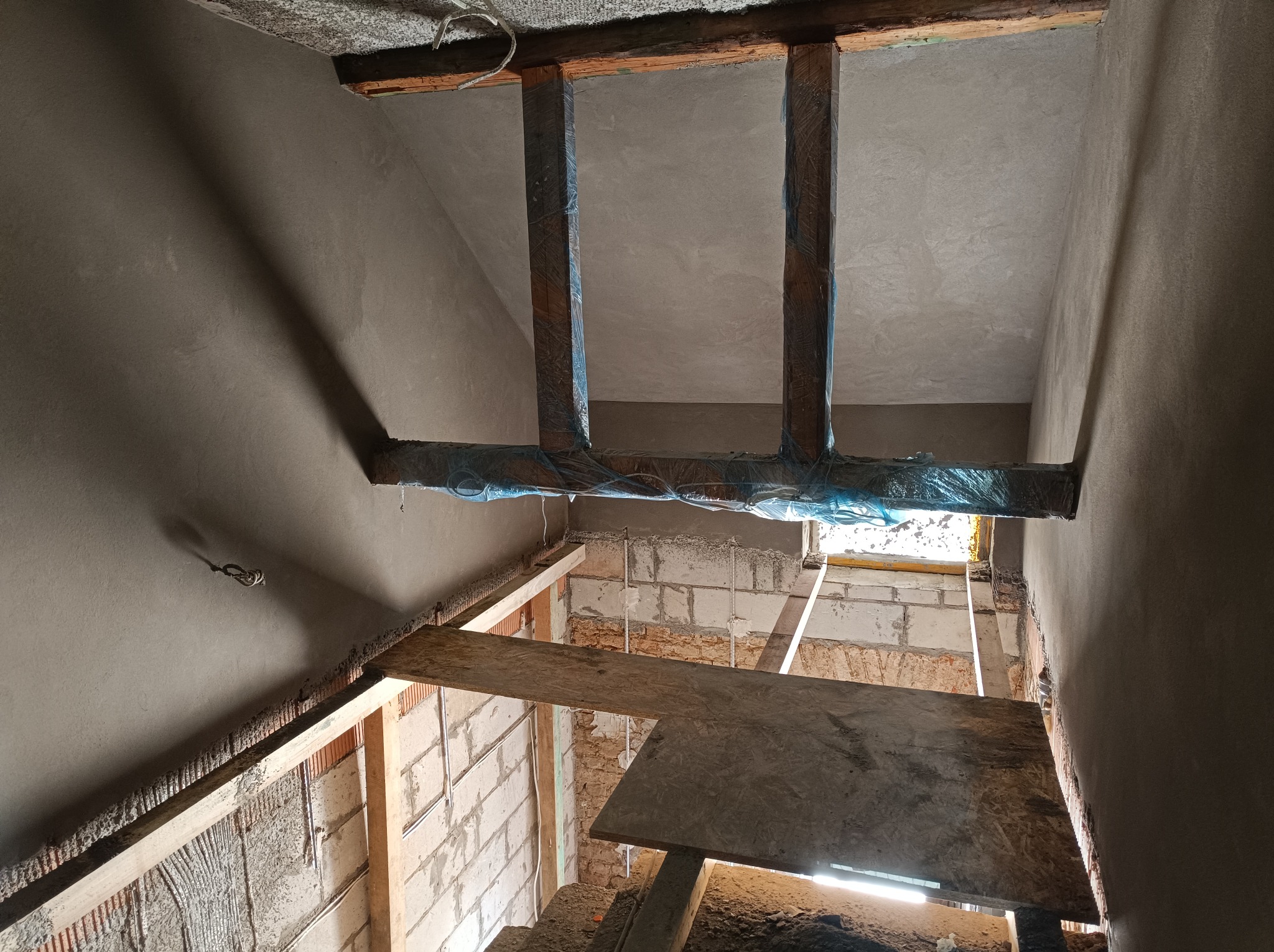 Surowe wnętrze w trakcie prac wykończeniowych; widoczne otynkowane ściany i sufit, drewniane belki zabezpieczone folią, fragment muru z cegieł i prowizoryczna drewniana kładka.