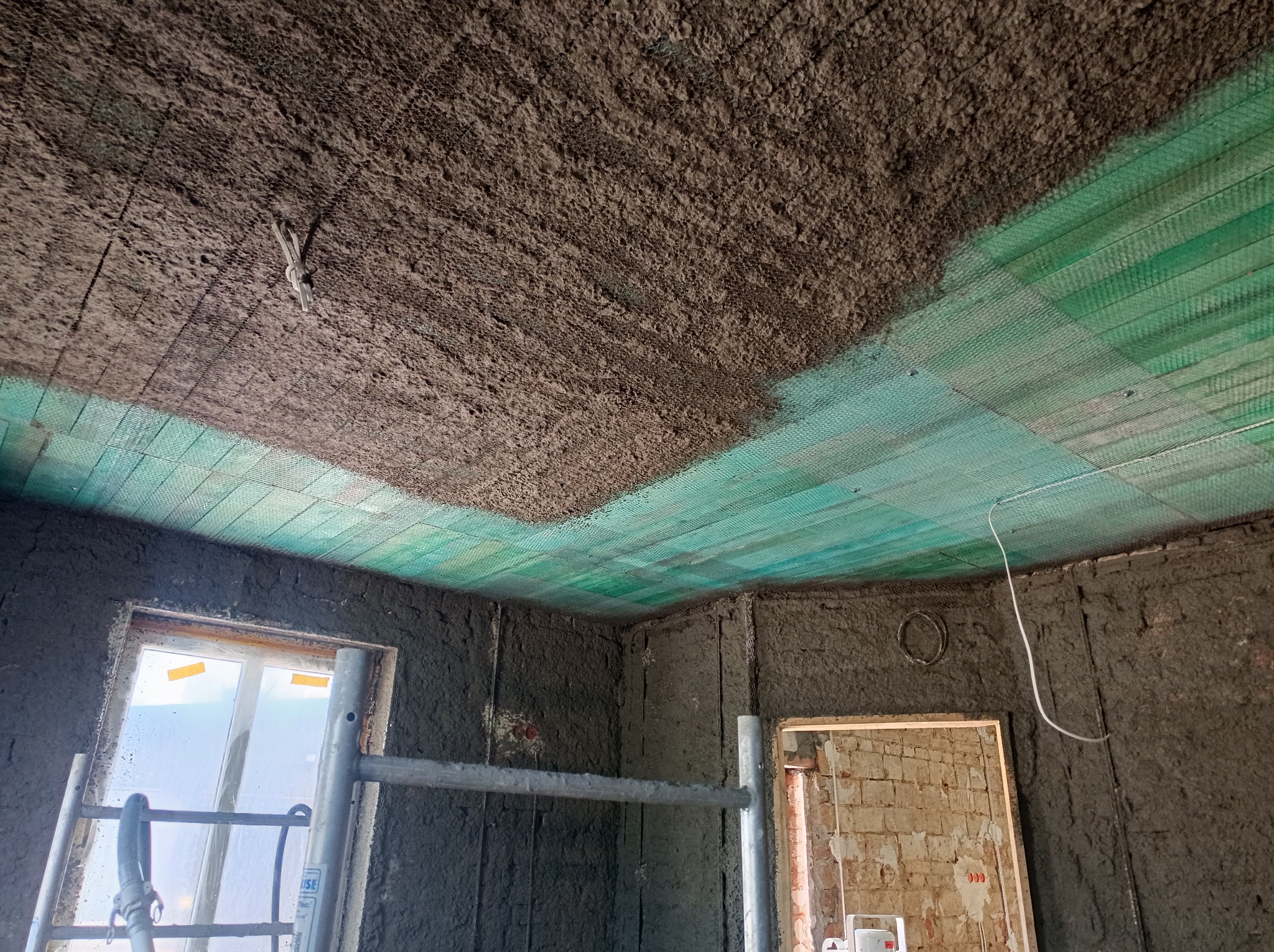 Wnętrze w trakcie tynkowania maszynowego: ściany i sufit pokryte szarym tynkiem, widoczna zielona siatka na suficie, fragment rusztowania przy oknie i otwór drzwiowy z widoczną cegłą.