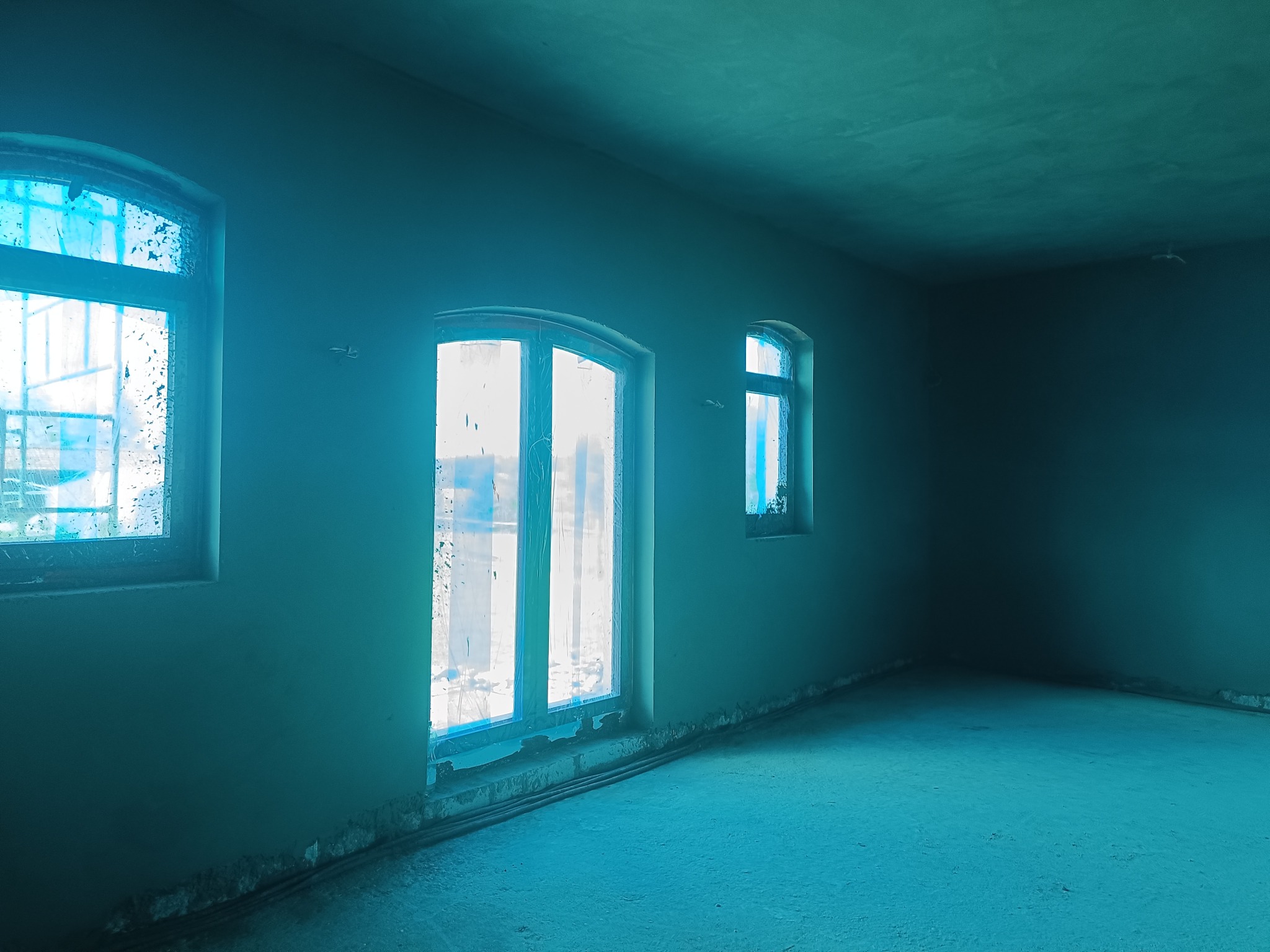 Wnętrze po tynkowaniu: trzy okna z folią ochronną, surowe ściany i podłoga w odcieniach turkusu, przygotowane do dalszych prac wykończeniowych. Widok na róg pomieszczenia.