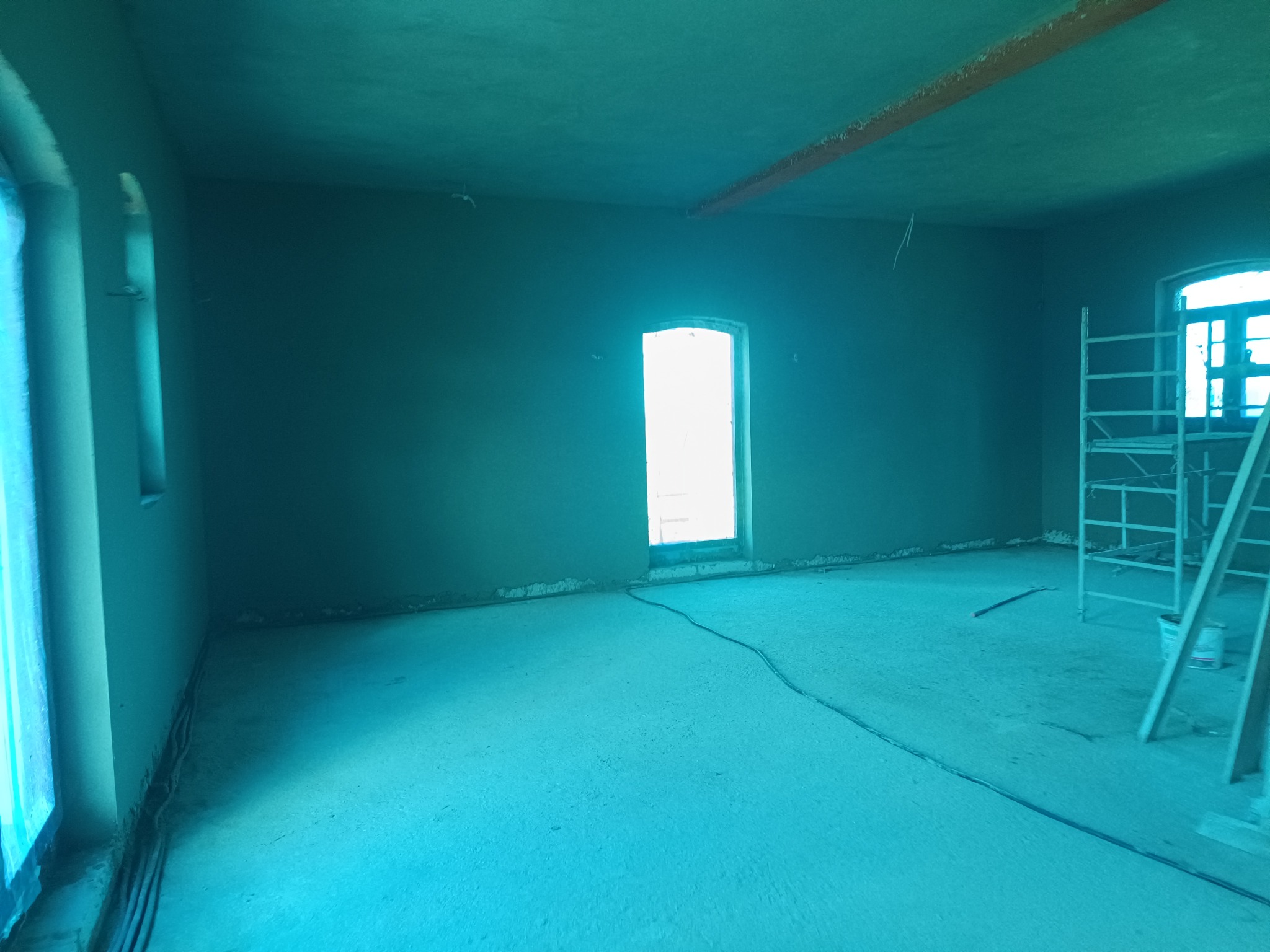 Wnętrze po nałożeniu tynku maszynowego: ściany świeżo pokryte, widoczne okna i rusztowanie. Surowy stan wykończenia, przygotowany do dalszych prac.