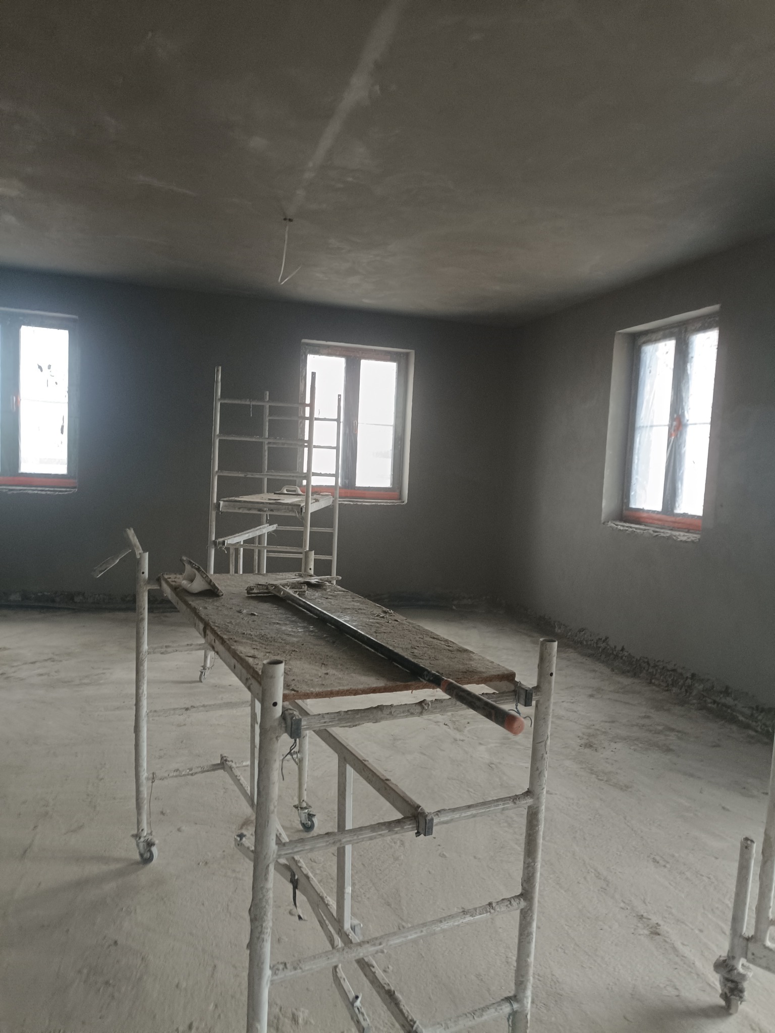 Pokój po tynkowaniu maszynowym: ściany i sufit pokryte świeżym tynkiem, widoczne rusztowania i narzędzia. Surowy stan wykończenia, wnętrze z trzema oknami.