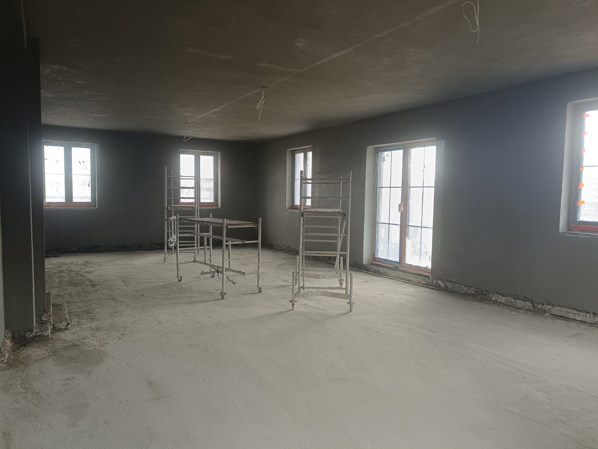 Wnętrze po tynkowaniu maszynowym: szare ściany, sufit i podłoga, widoczne rusztowania na kółkach, okna z widokiem na zewnątrz. Minimalistyczny, surowy stan wykończenia.