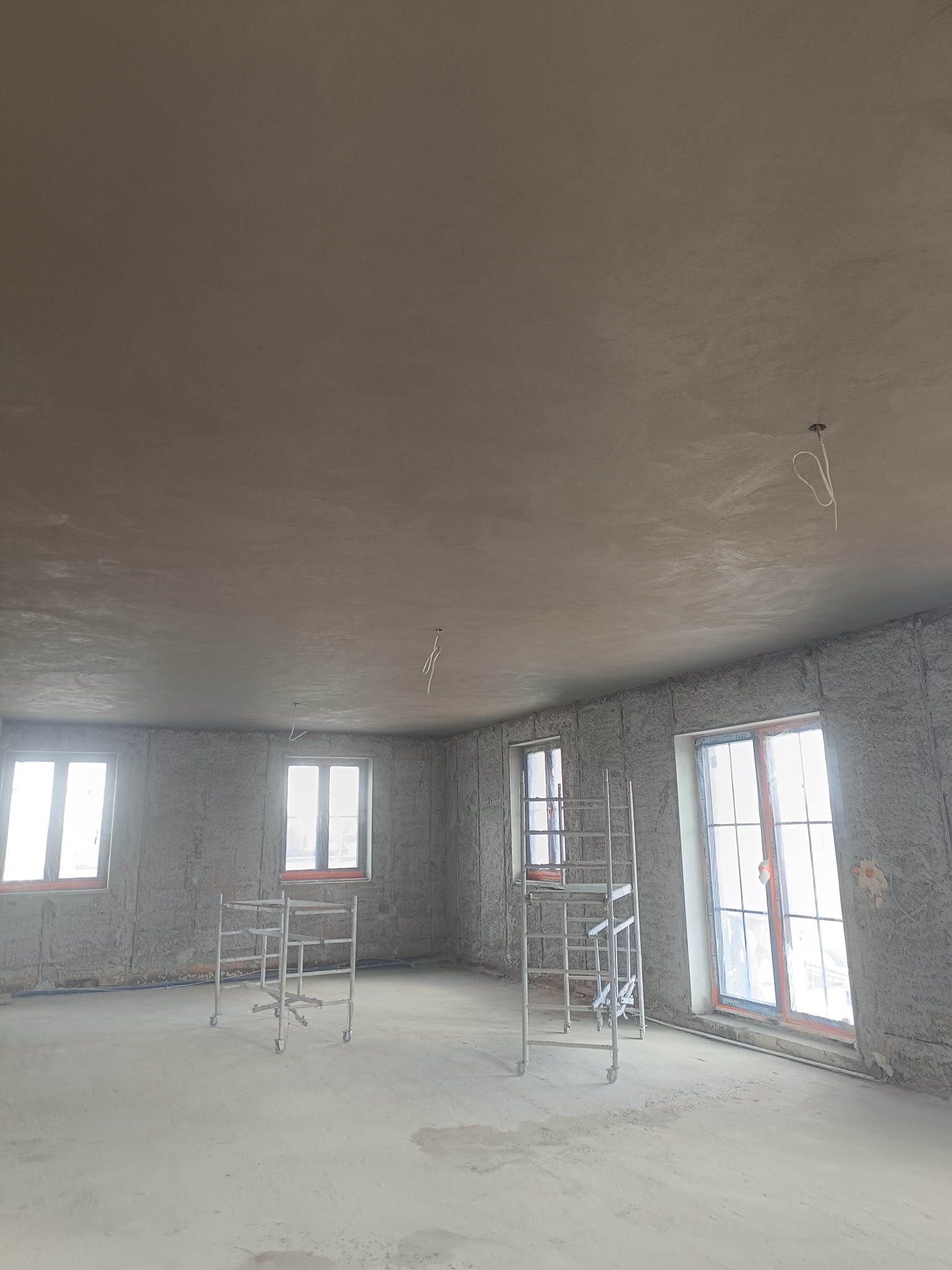 Wnętrze w trakcie prac wykończeniowych: ściany pokryte surowym tynkiem, metalowe rusztowania przy oknach. Widoczne przewody elektryczne na suficie i niebieska rurka przy podłodze.