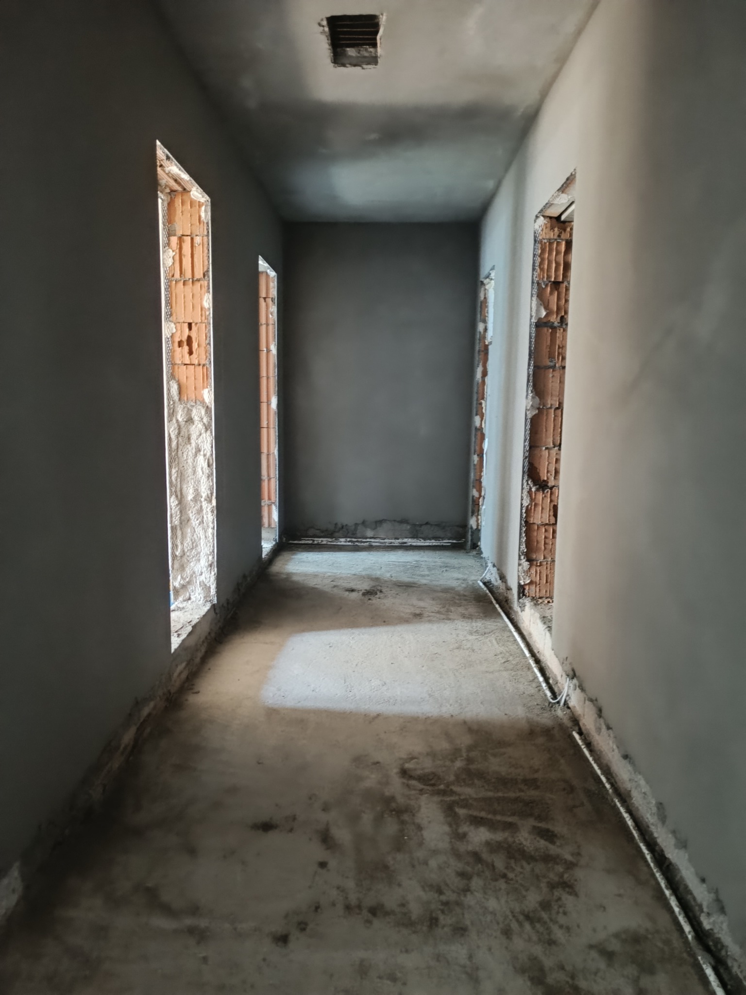 Wnętrze korytarza w trakcie prac wykończeniowych: ściany po tynkowaniu, widoczne fragmenty cegieł w otworach drzwiowych, surowa posadzka.
