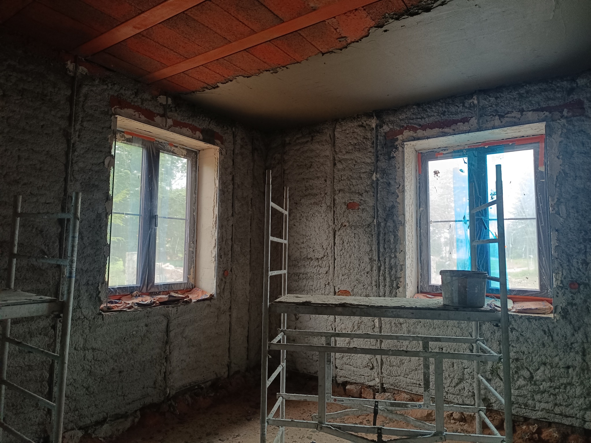 Wnętrze w trakcie prac tynkarskich: ściany pokryte surowym tynkiem, widoczne rusztowania i okna zabezpieczone folią. Fragment sufitu z ceglanym wykończeniem.