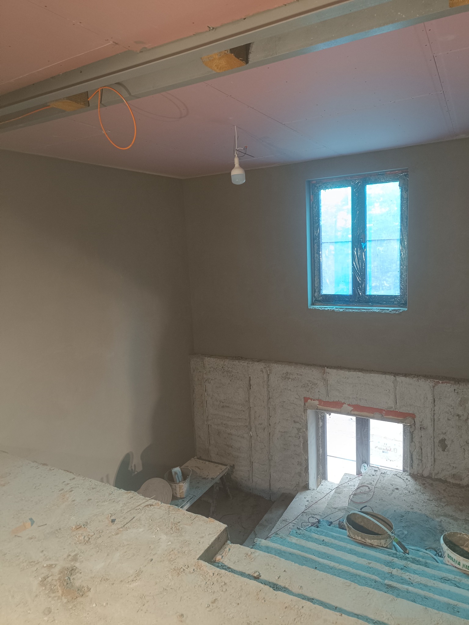 Wnętrze w trakcie prac wykończeniowych: ściany po tynkowaniu, okno zabezpieczone folią, widoczne narzędzia i materiały budowlane. Częściowo otynkowana ściana schodów.