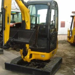 Żółta, kompaktowa maszyna budowlana JCB 8018 z rozsuwanymi gąsienicami, widoczna z przodu kabina, obok i w tle inne maszyny budowlane, na placu budowy.