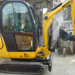 Żółta, gąsienicowa maszyna budowlana JCB 8018 z rozsuwanymi gąsienicami na tle budynku z cegły i wózka.