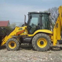 Żółta maszyna budowlana JCB 3CX Contractor z łyżką z przodu i ramieniem kopiącym z tyłu, stojąca na żwirowym terenie, w tle widoczny budynek mieszkalny i furgonetka serwisowa JCB.